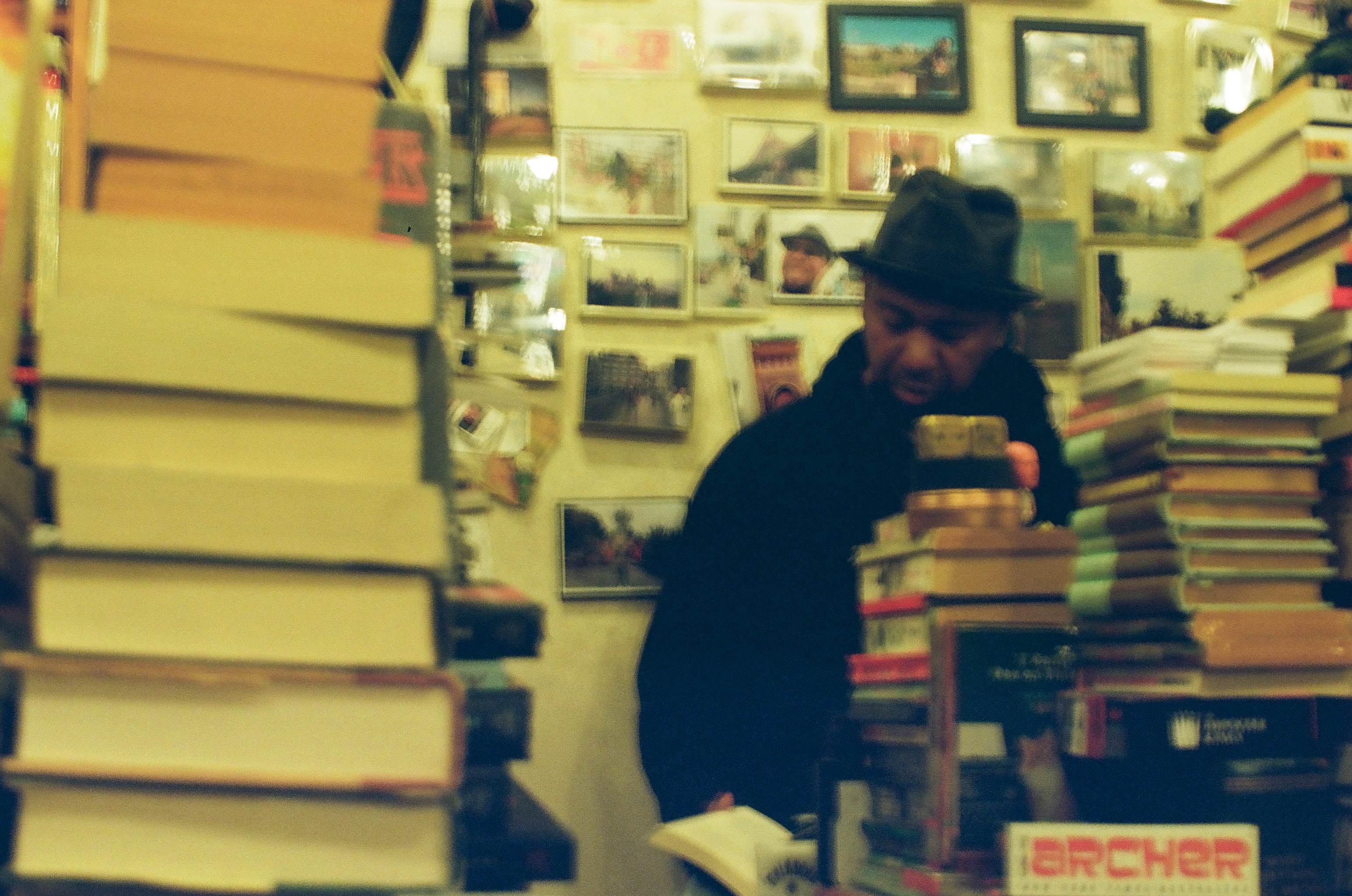 A man leans behind tall stacks of books beneath walls densely covered with framed pictures and prints.