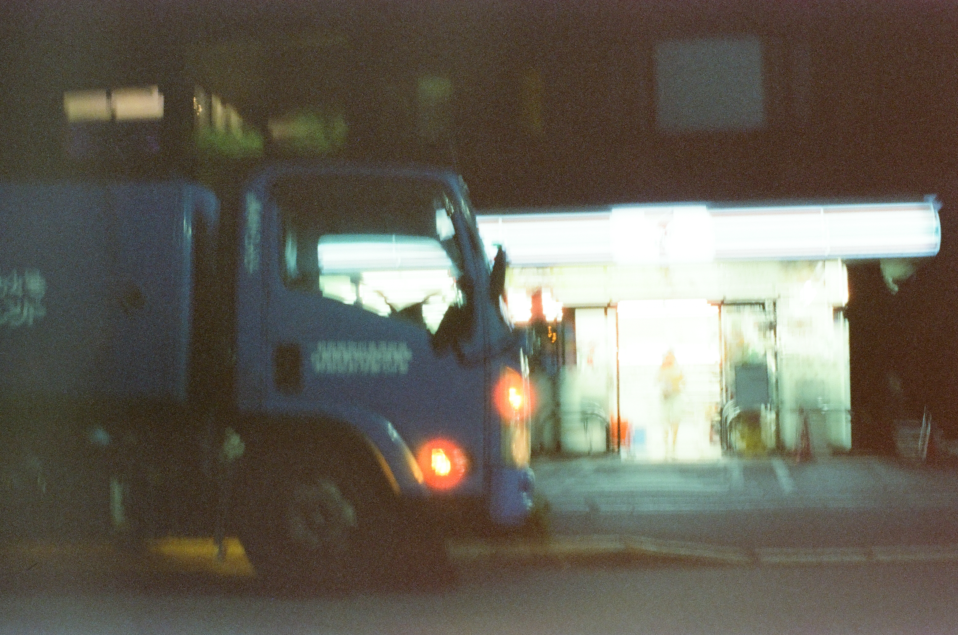 A blue box truck is parked in the foreground before an illuminated convenience store where a figure stands near the glass entrance.