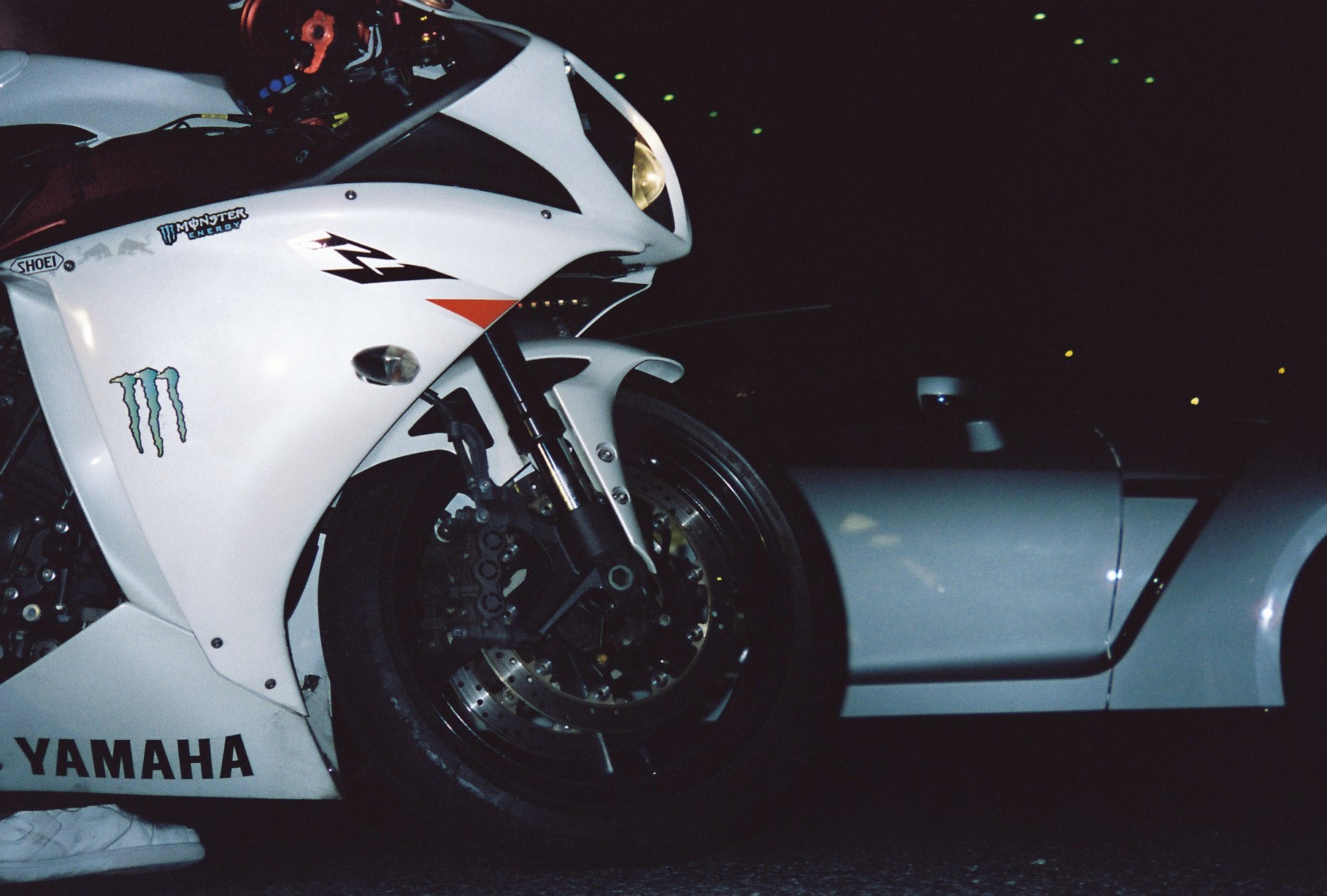 Low view of a white motorcycle's front wheel, forks, and fairing beside the pale rear fender of a car in darkness.
