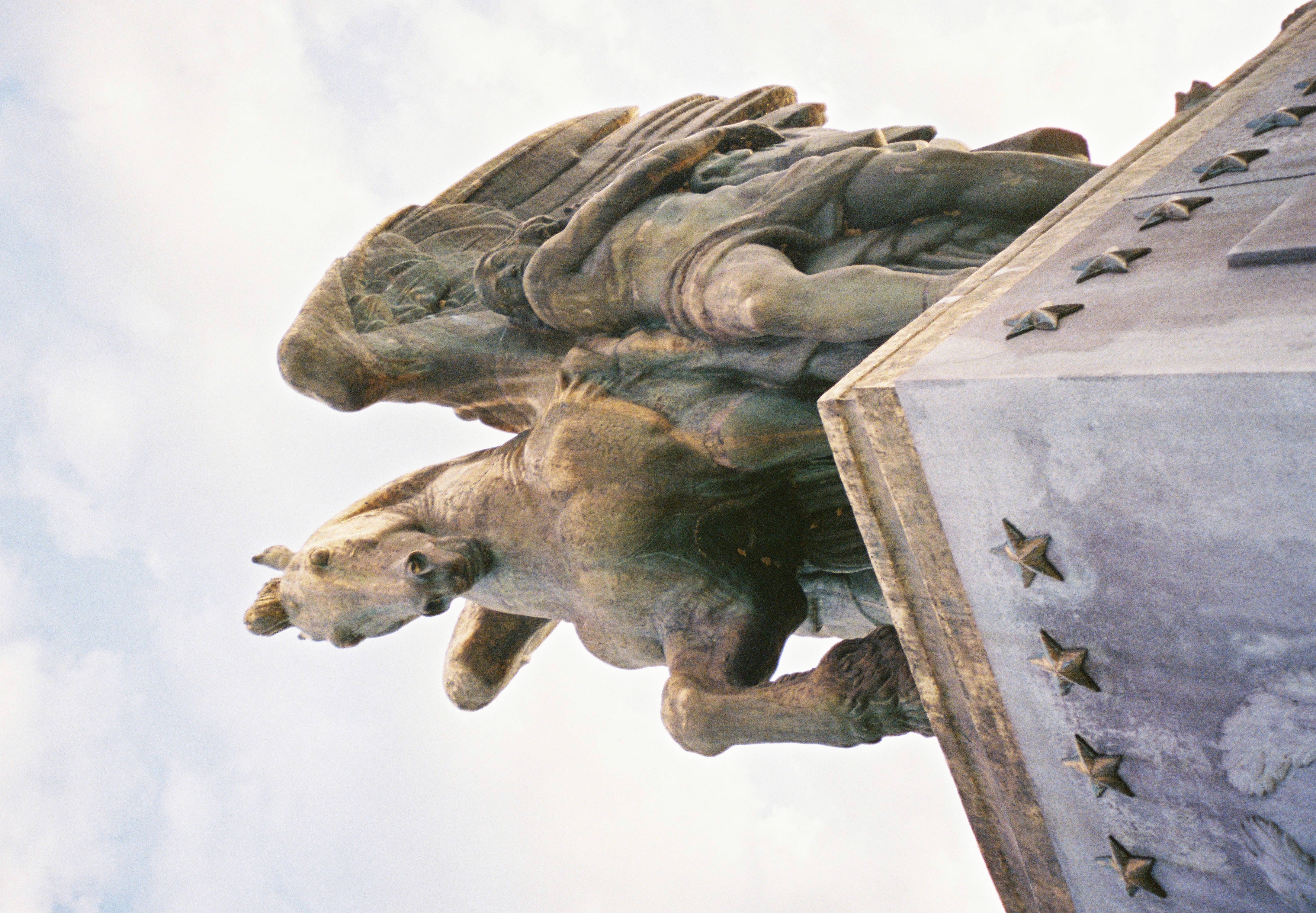 Low-angle view of a green-patinated winged horse rearing beside a mostly unclothed human figure with drapery around the lower body, atop a stone pedestal edged with raised stars.