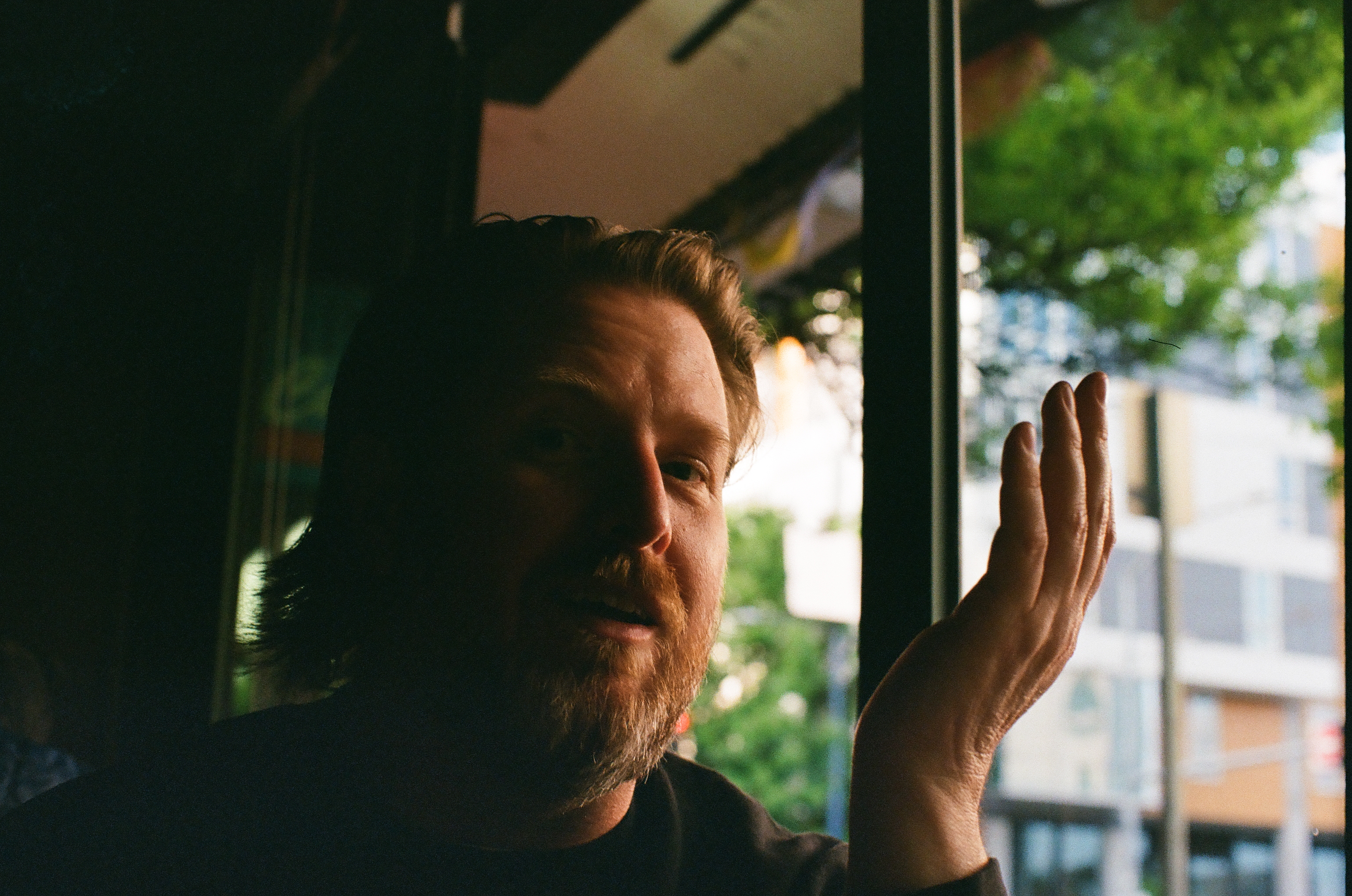 A bearded person raises one hand beside a window with green trees and buildings outside.