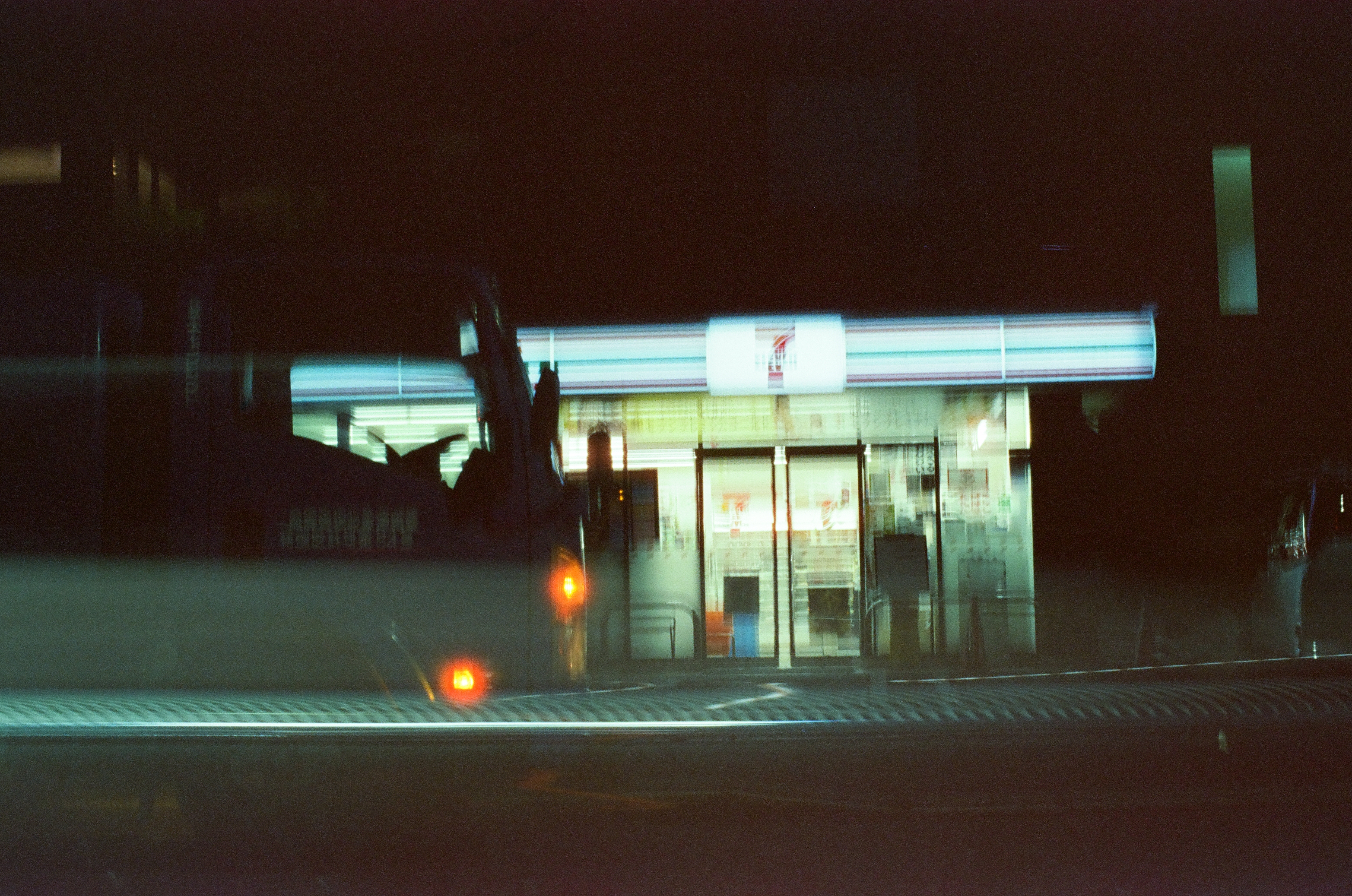 An illuminated convenience store canopy displays a bold numeral 7 above glass doors, seen across an empty dark street with a small orange light.