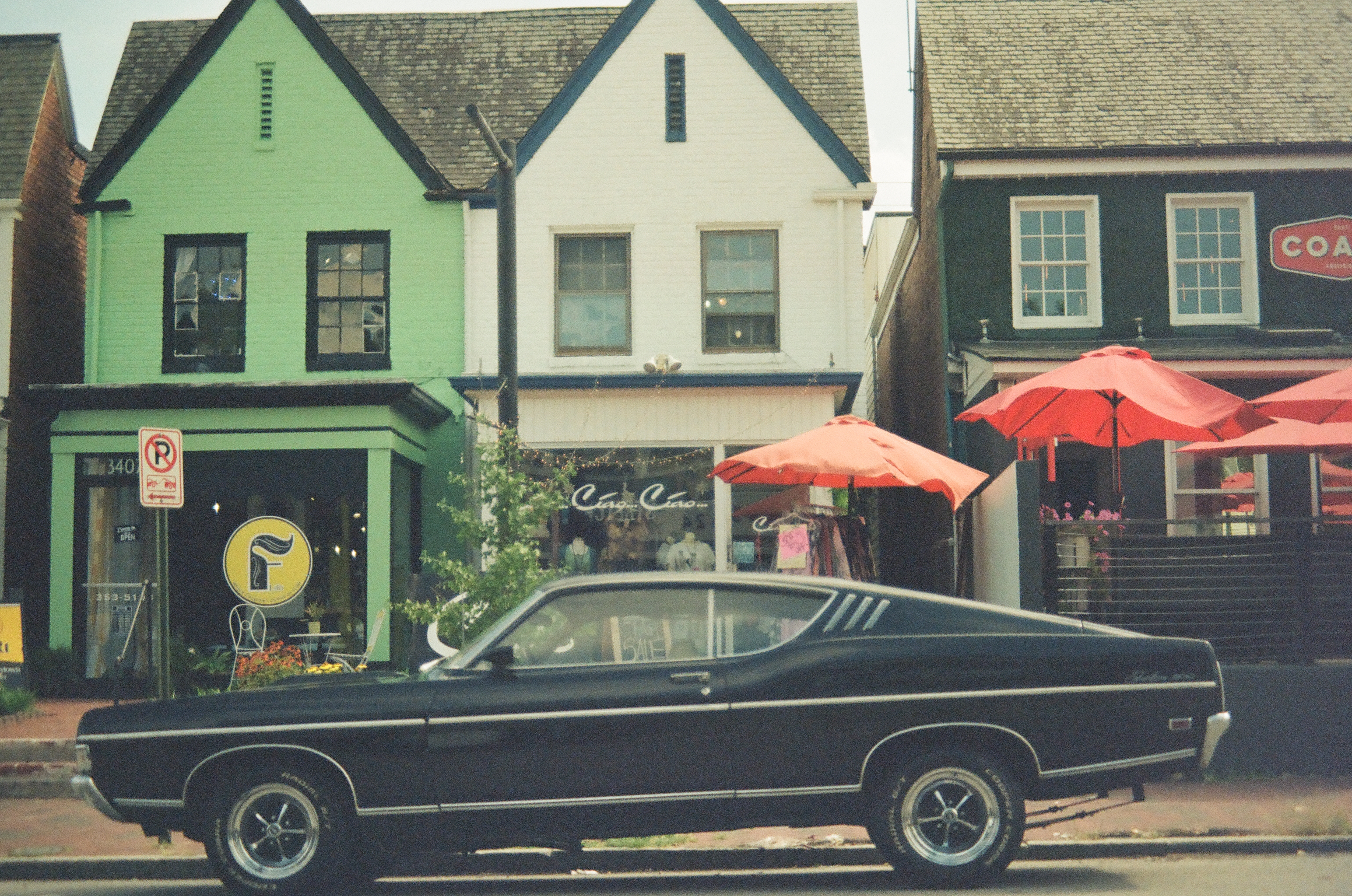 A black vintage fastback car is parked along a curb in front of green, white, and dark storefronts under red patio umbrellas.