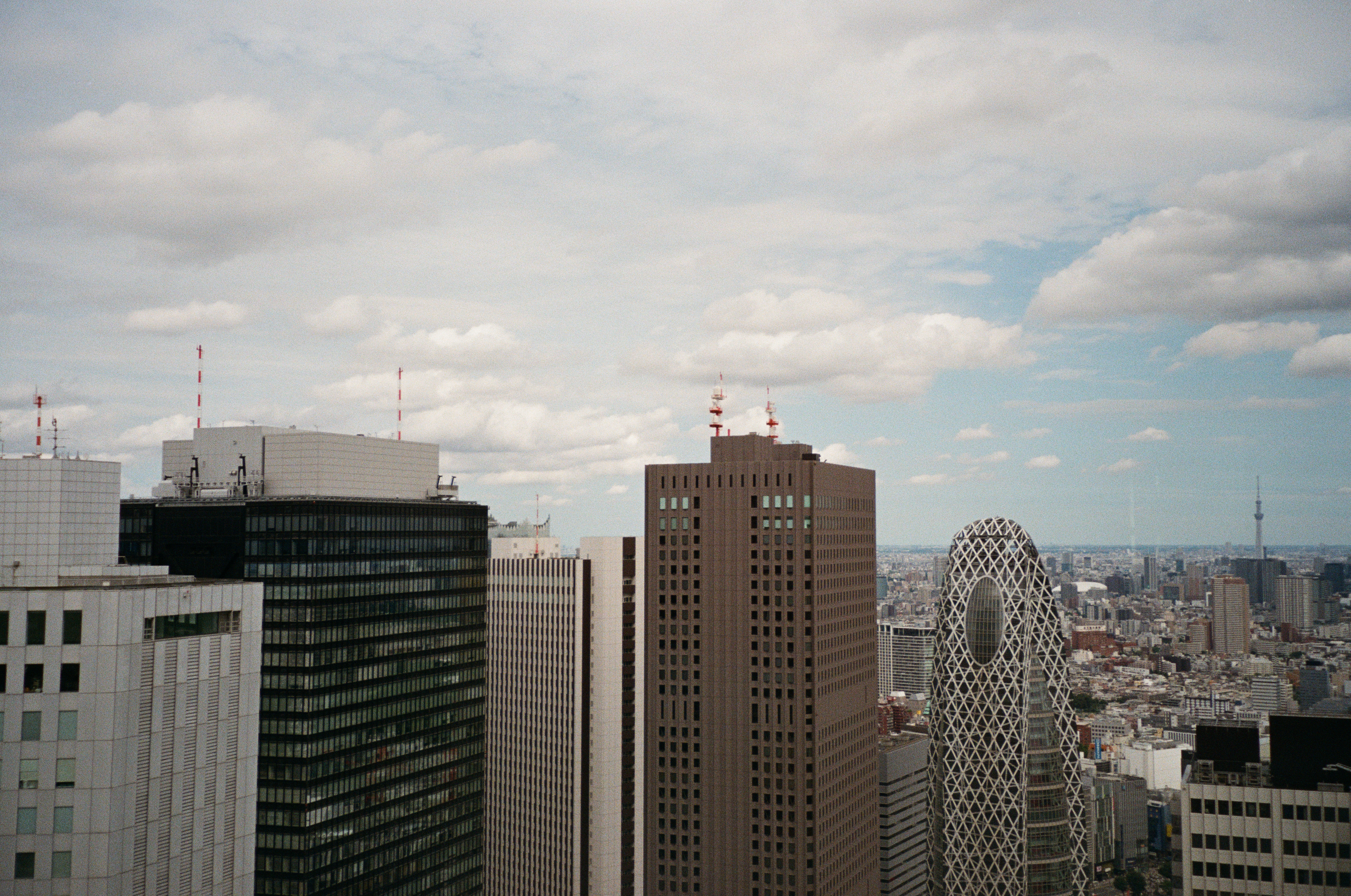 A row of high-rise towers, including one with a diamond-lattice facade and an oval opening, stands beneath blue sky and fat cumulus clouds.