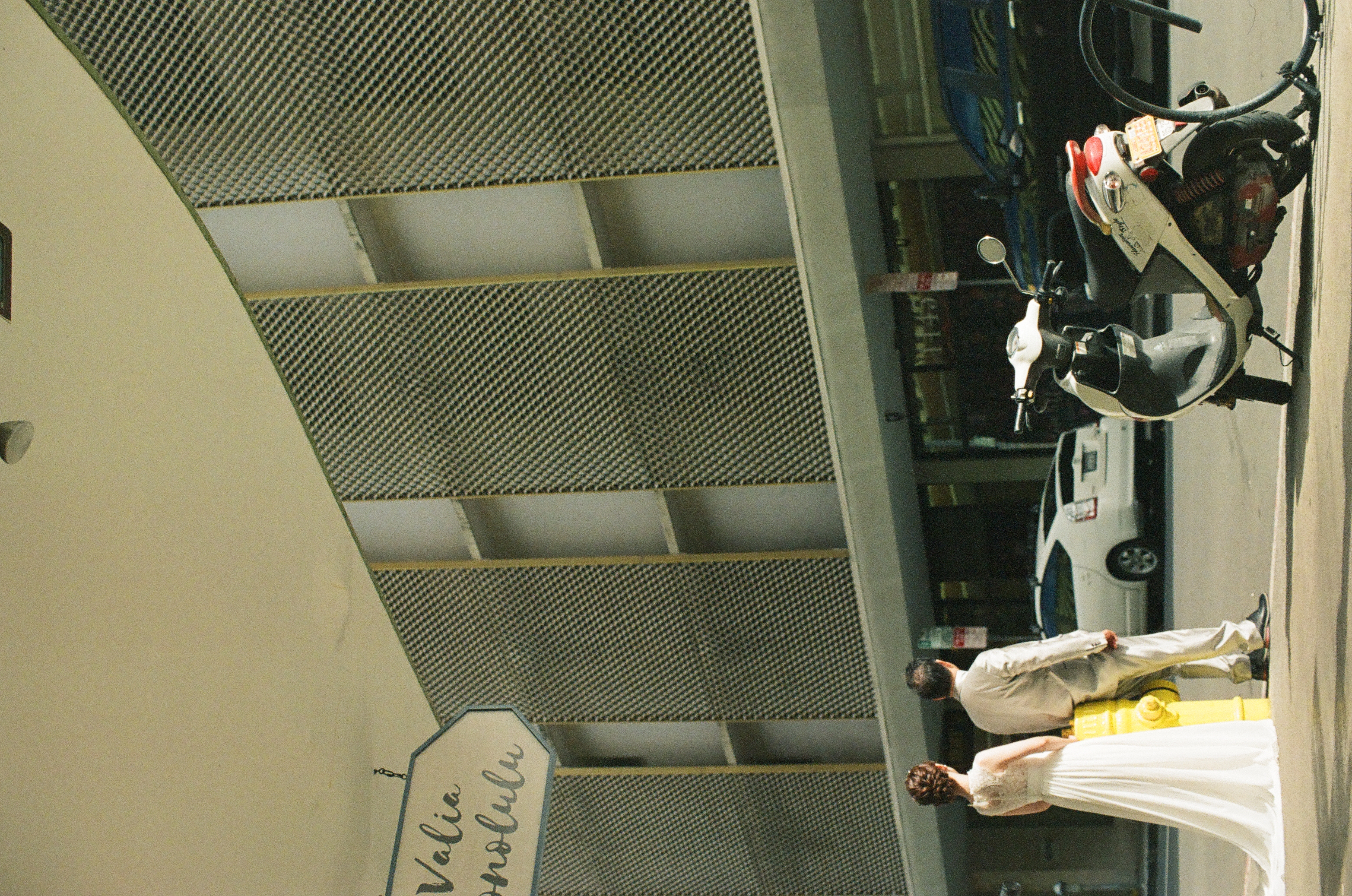 Two people in long pale formal clothes stand near a parked scooter outside a modern building.