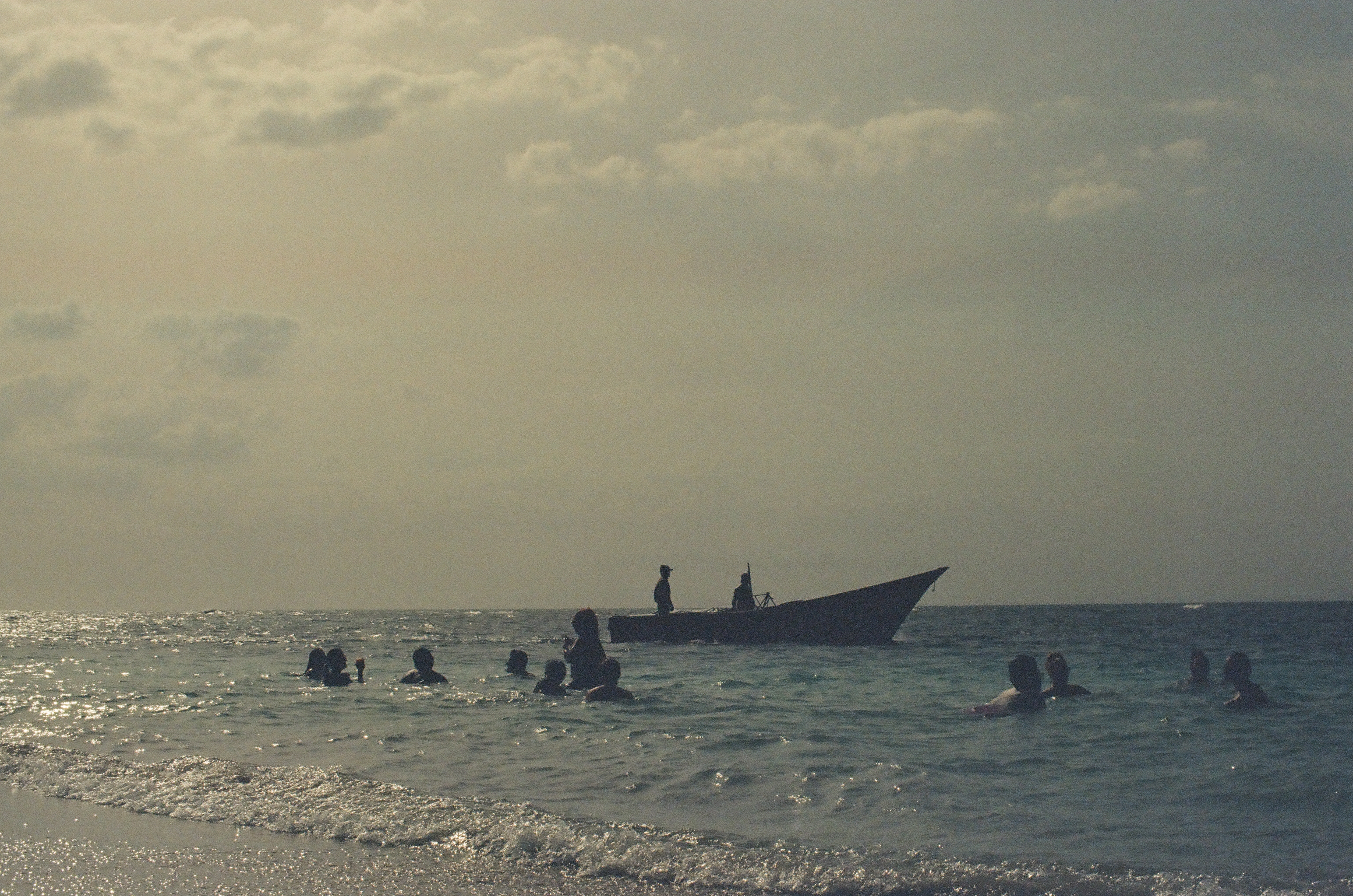 A cluster of silhouetted swimmers bob in calm ocean water beside a small boat where two figures stand, beneath a hazy overcast sky.