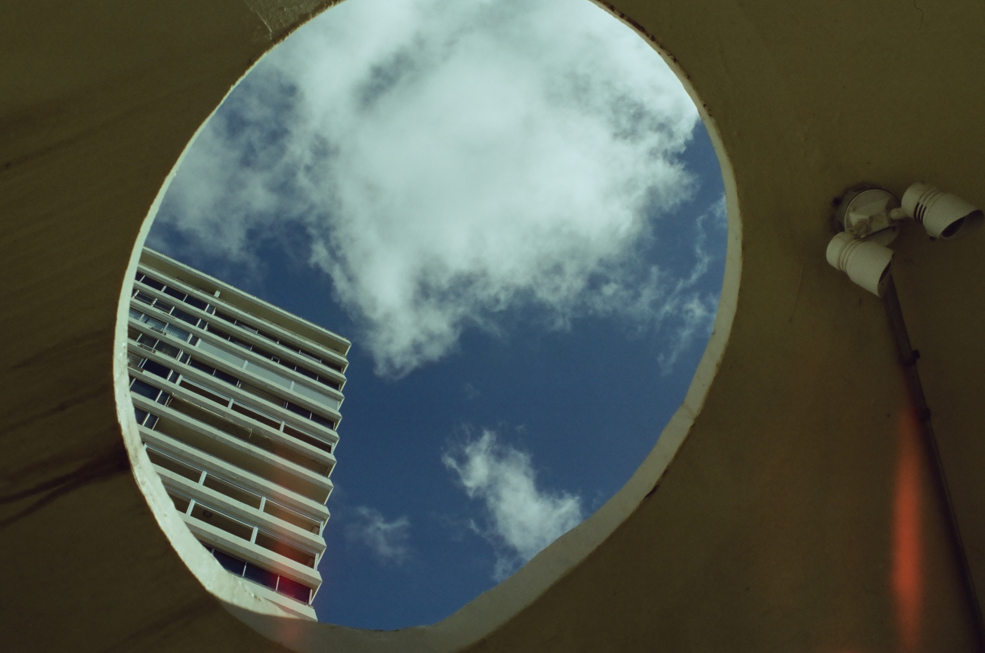 A round concrete opening frames blue sky, a puffy white cloud, and the corner of a balconied tower, with a wall lamp just outside its edge.