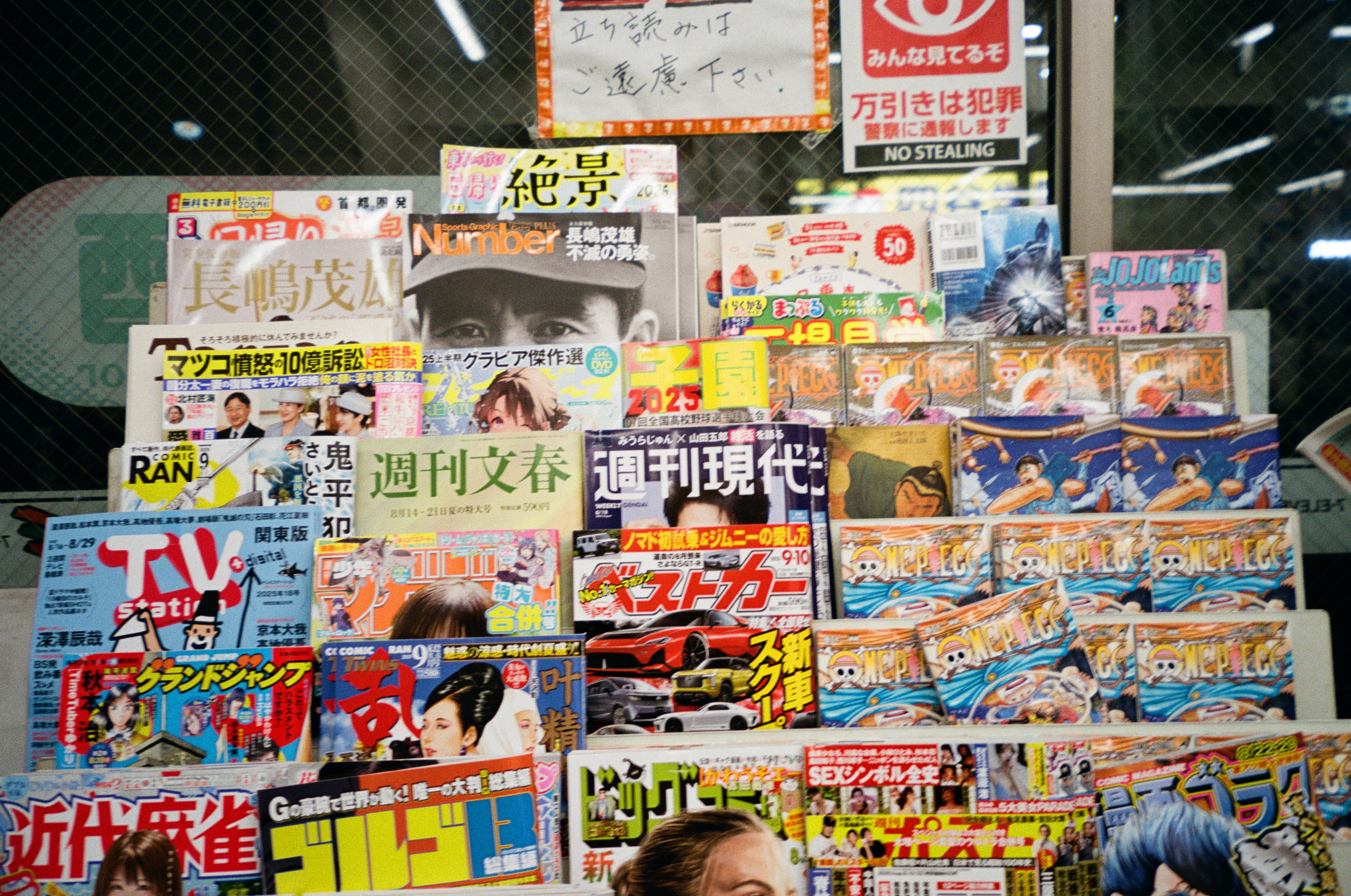 A magazine rack packed with Japanese-language weeklies, manga, and comics sits below signs reading NO STEALING and warning against shoplifting.