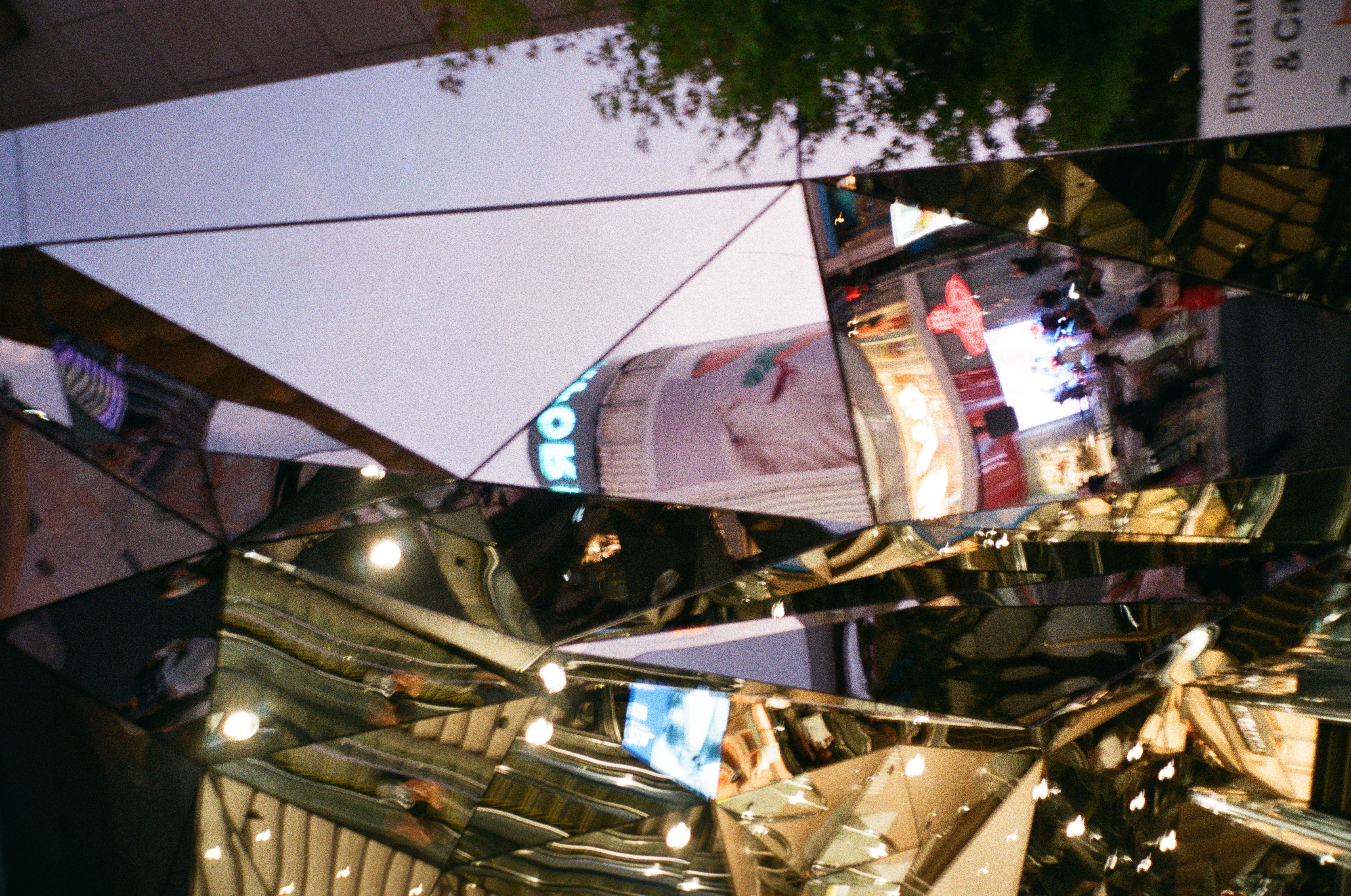 A tilted, faceted mirror facade splits reflections of illuminated signage, tree canopy, and string lights into angular fragments at night.
