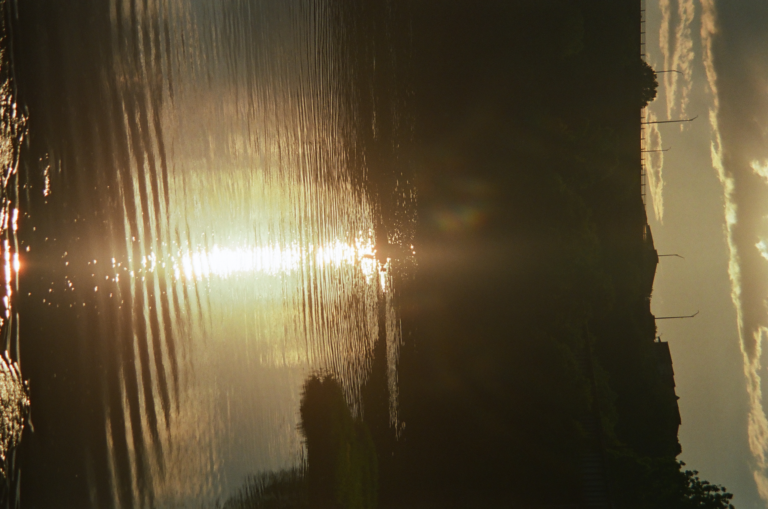 A bright column of reflected sunset light runs down rippled river water beneath a dark silhouetted bank and buildings.