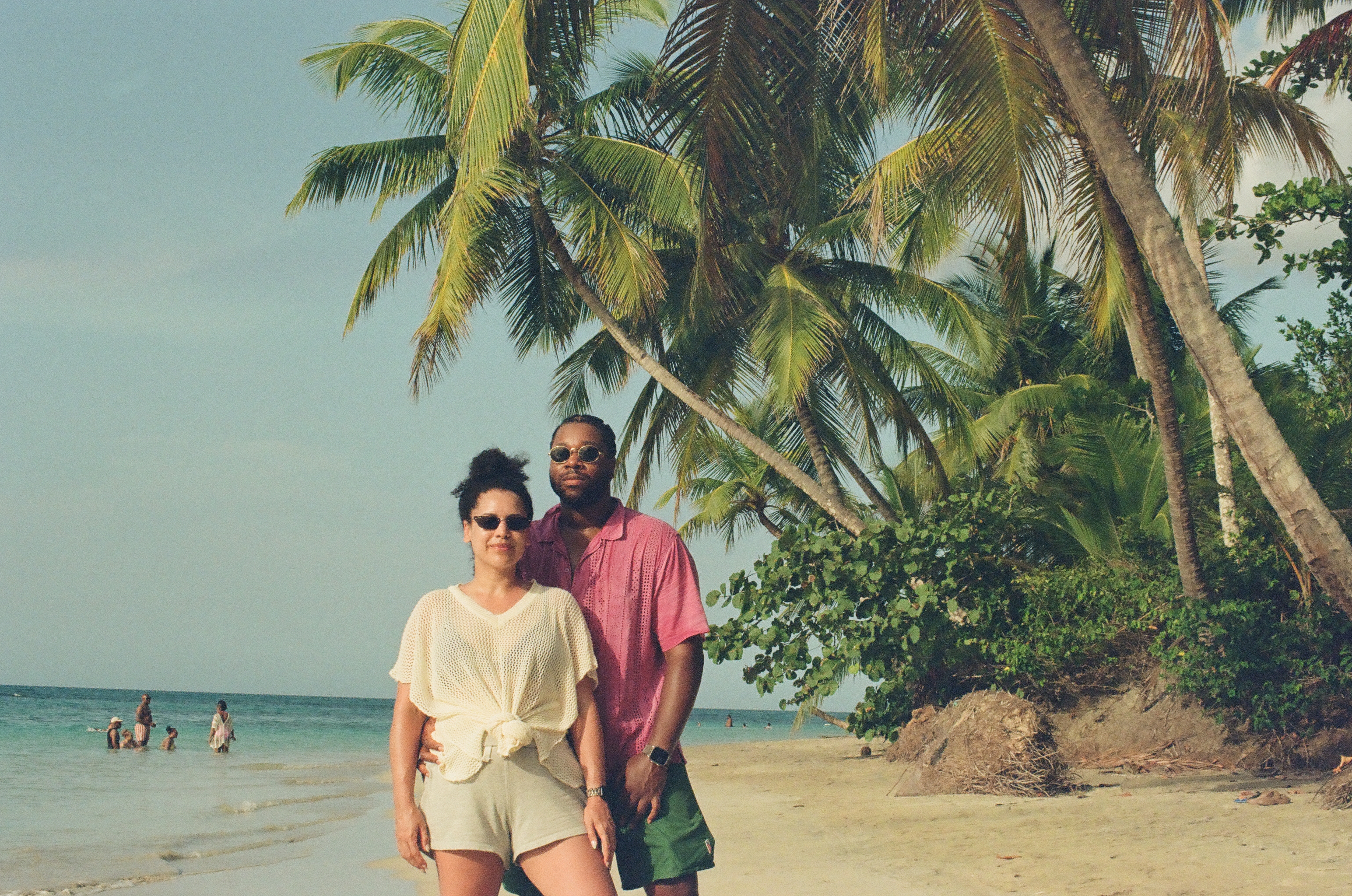 A woman in a white mesh top and a man in a pink shirt and green shorts stand together on sand before coconut palms and blue water.