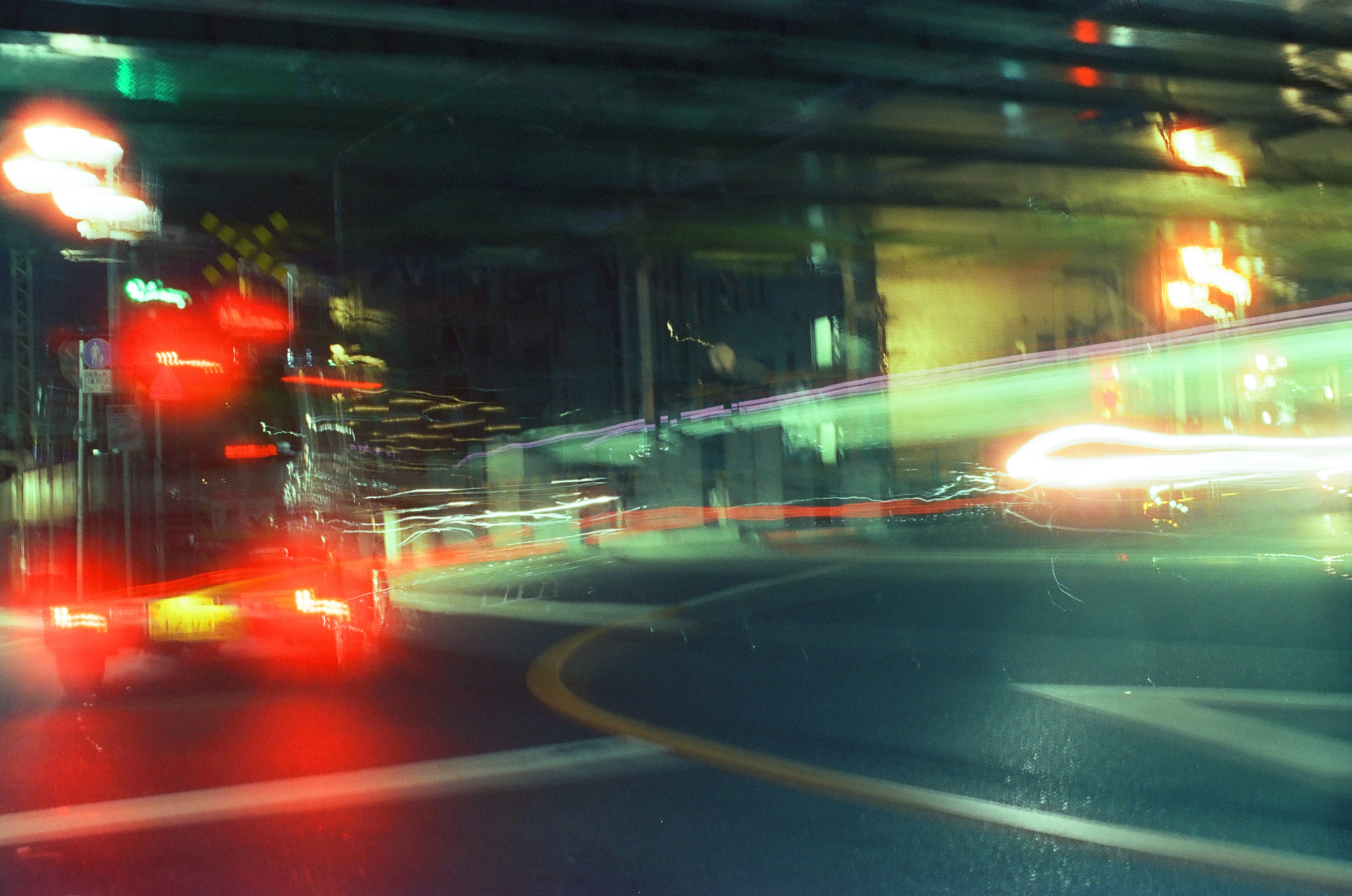 Red brake lights smear into curved ribbons at a night intersection while a pale green light trail and white streaks cross above the roadway.
