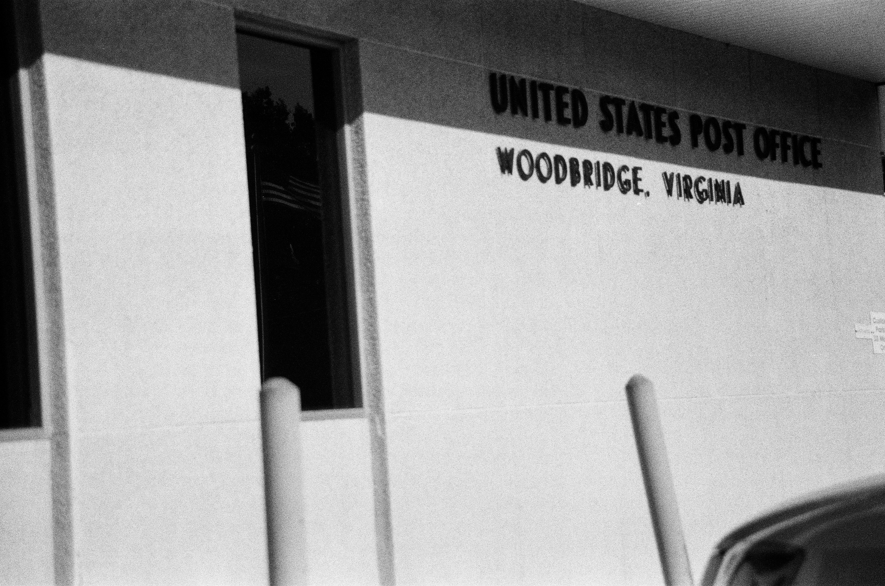 United States Post Office lettering spans a pale wall above upright posts and a cropped vehicle.
