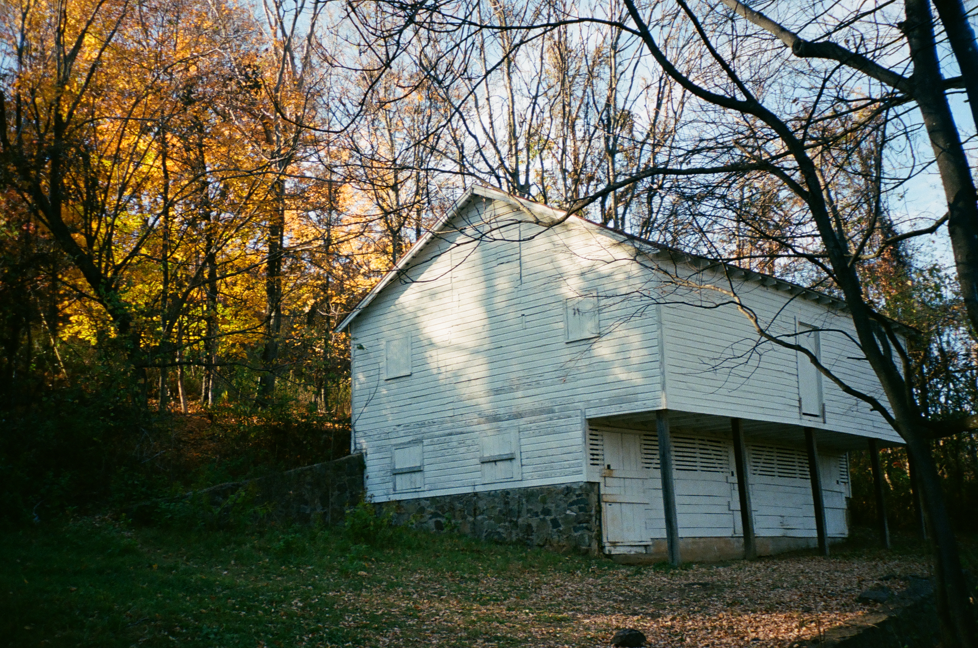 A large two-story white barn on a stone foundation stands among bare trees and fallen autumn leaves.