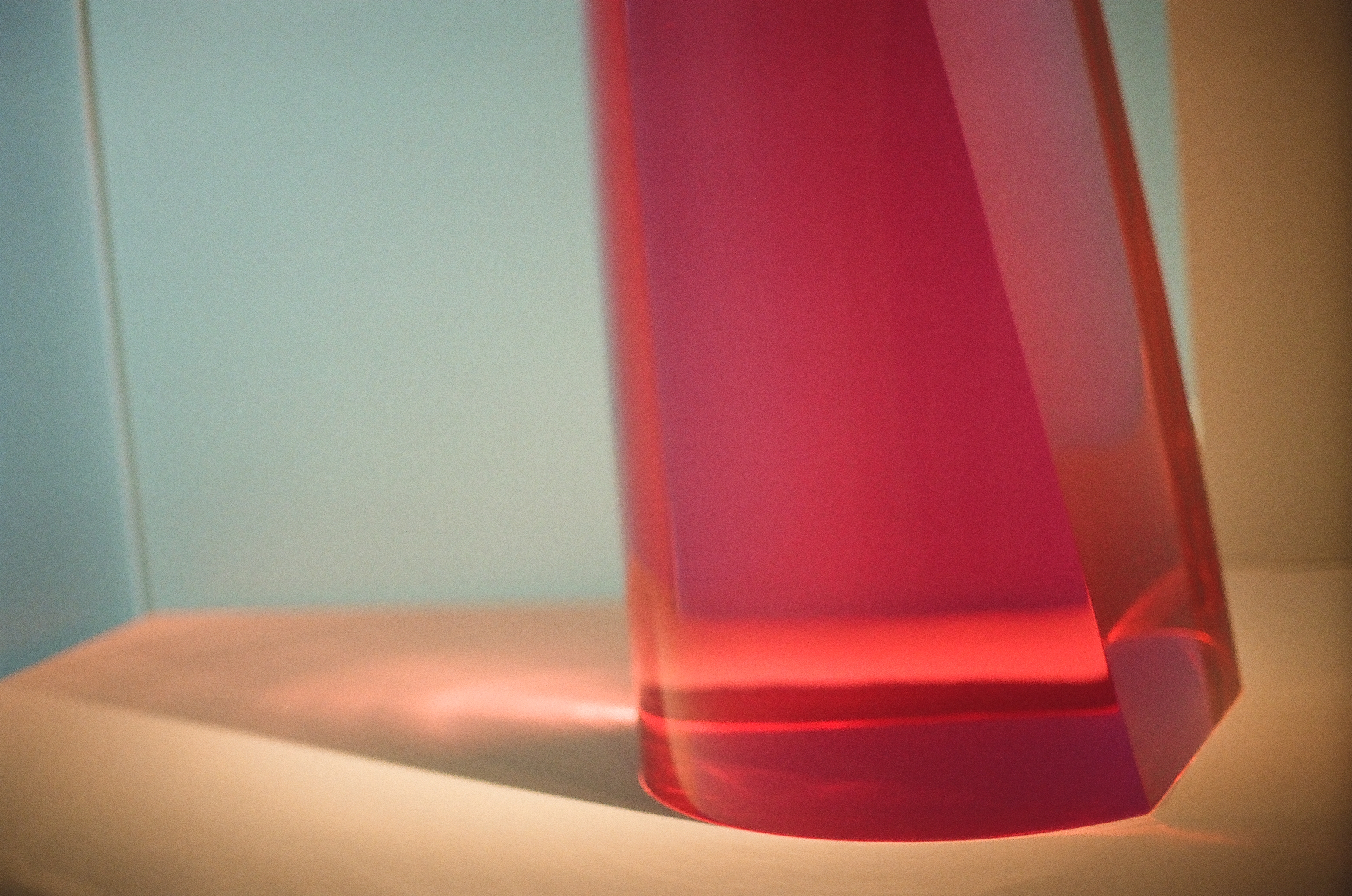 A translucent red tapering form stands on a cream surface against a pale blue wall, with rose light pooling at its base.