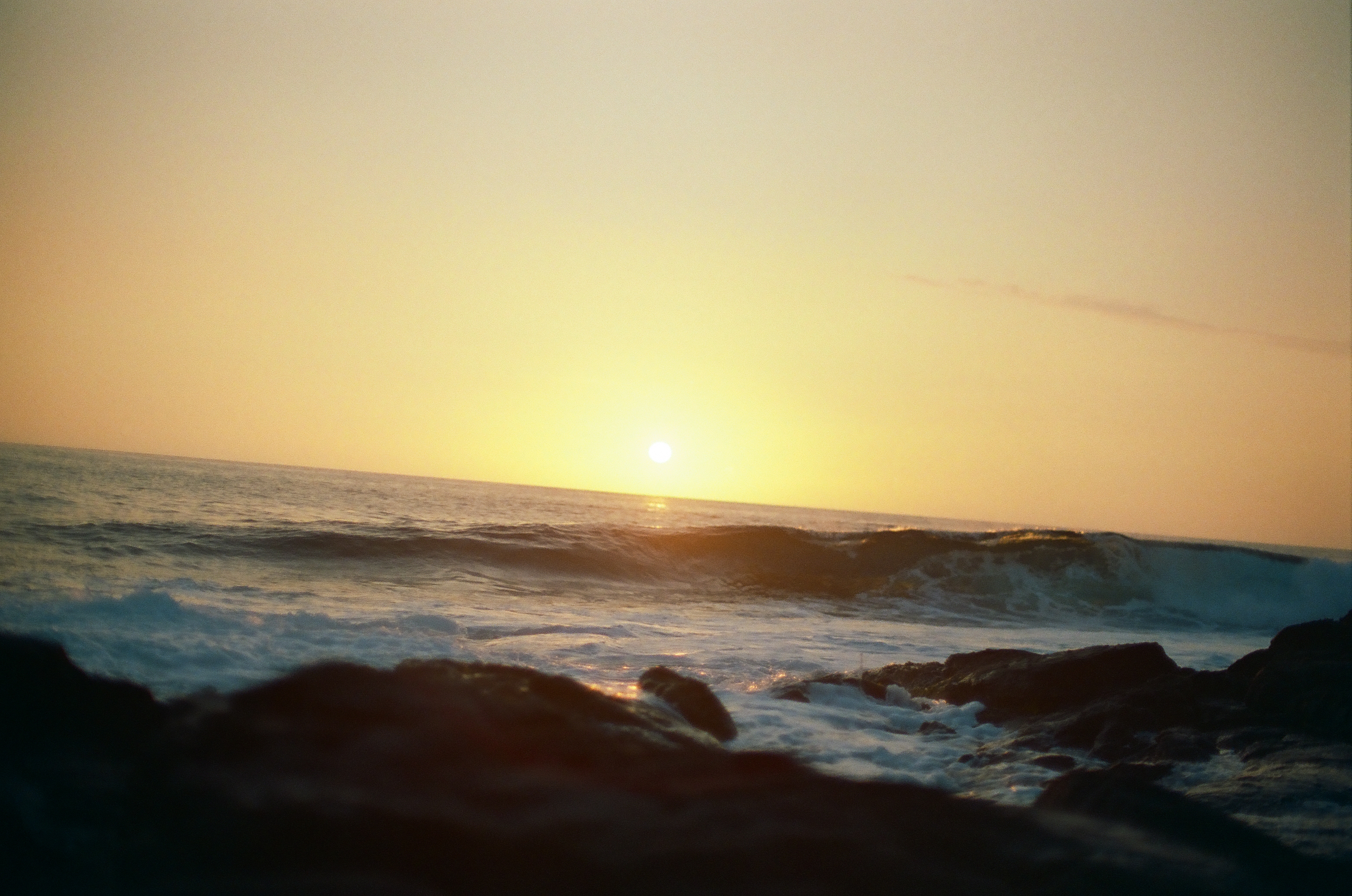 The low sun hangs above a curling wave that breaks through scattered dark rocks under a yellow sky.