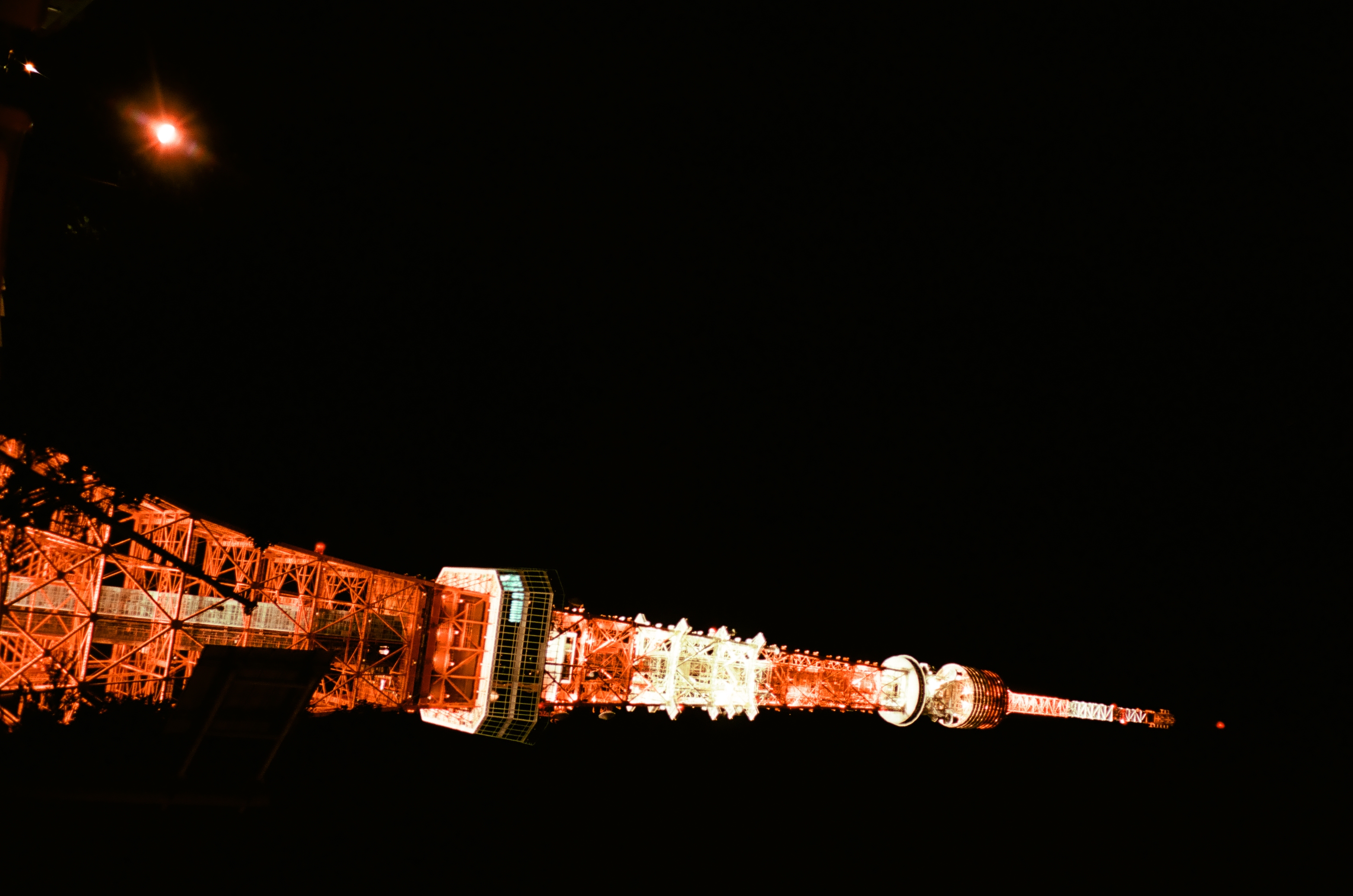 A tall red-and-white lattice tower tilts diagonally against a black night sky, with a lone streetlamp flaring at lower left.
