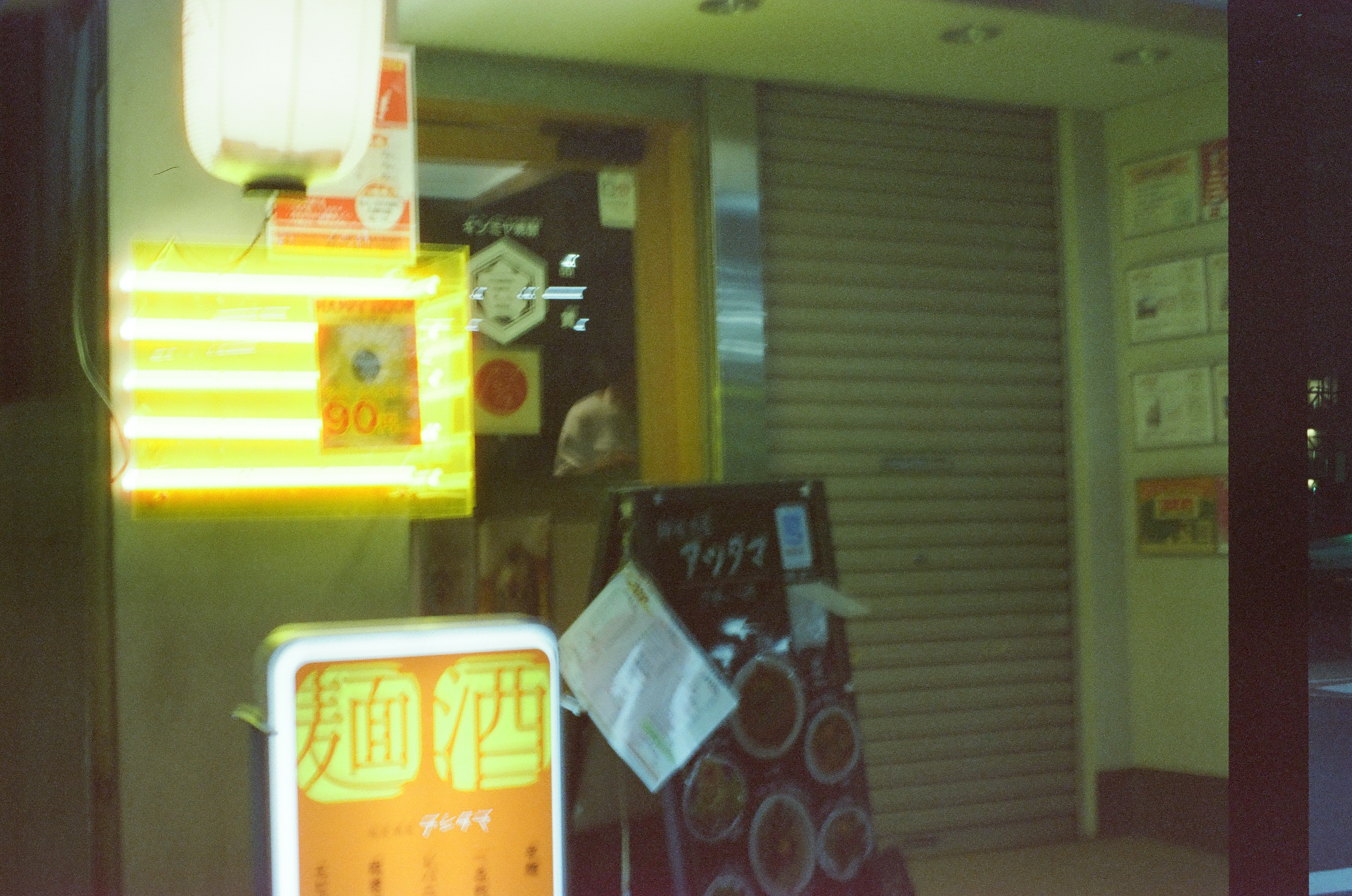 Illuminated signs cluster before a closed storefront and a dark rolled metal shutter at night.
