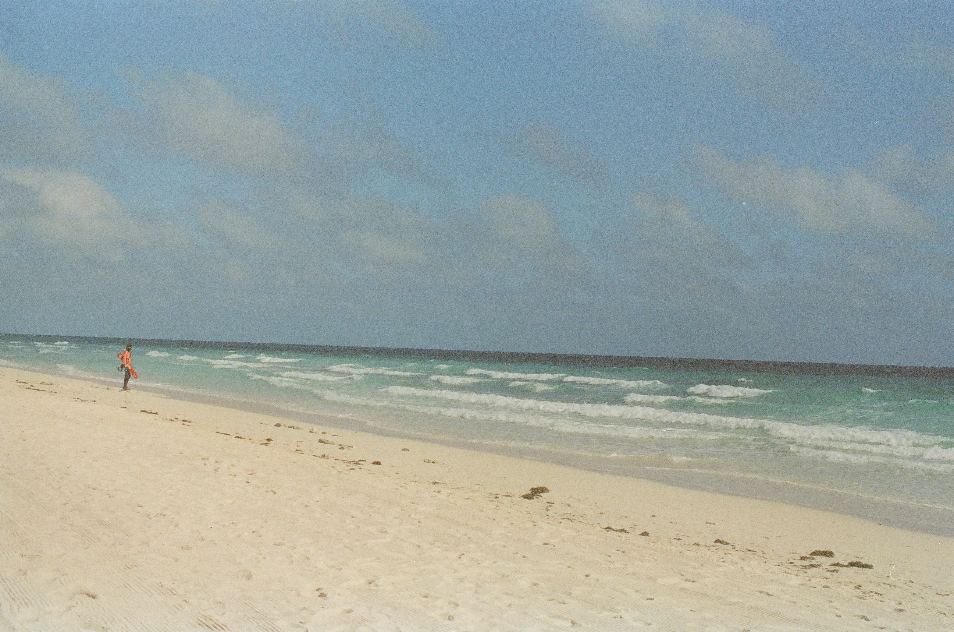 Pale sand marked with track lines stretches toward rows of breaking green surf, where a lone distant figure in an orange top stands near the water.