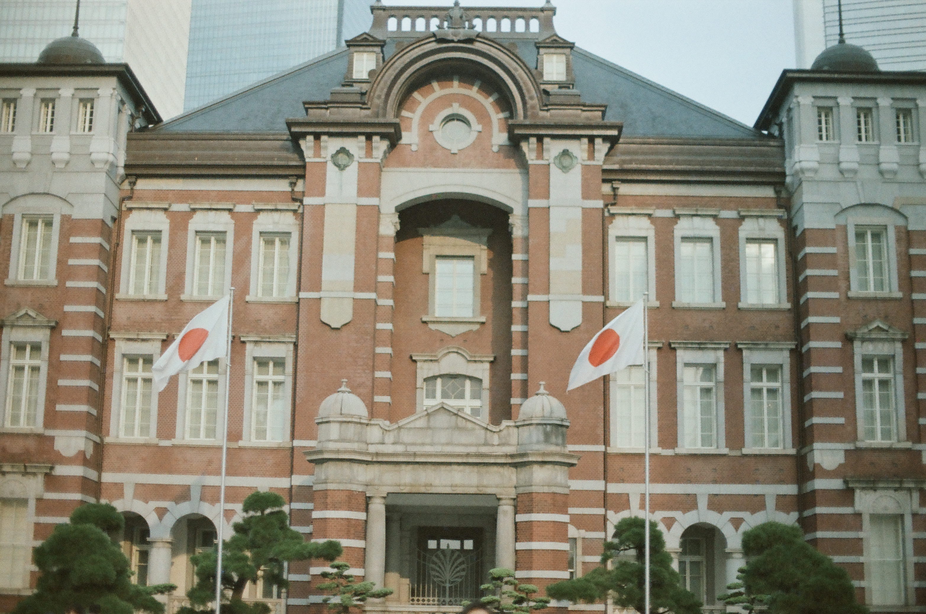 A red-brick building's ornate facade with arched windows flies two white flags, each with a red circular disc, above sculpted pine trees.