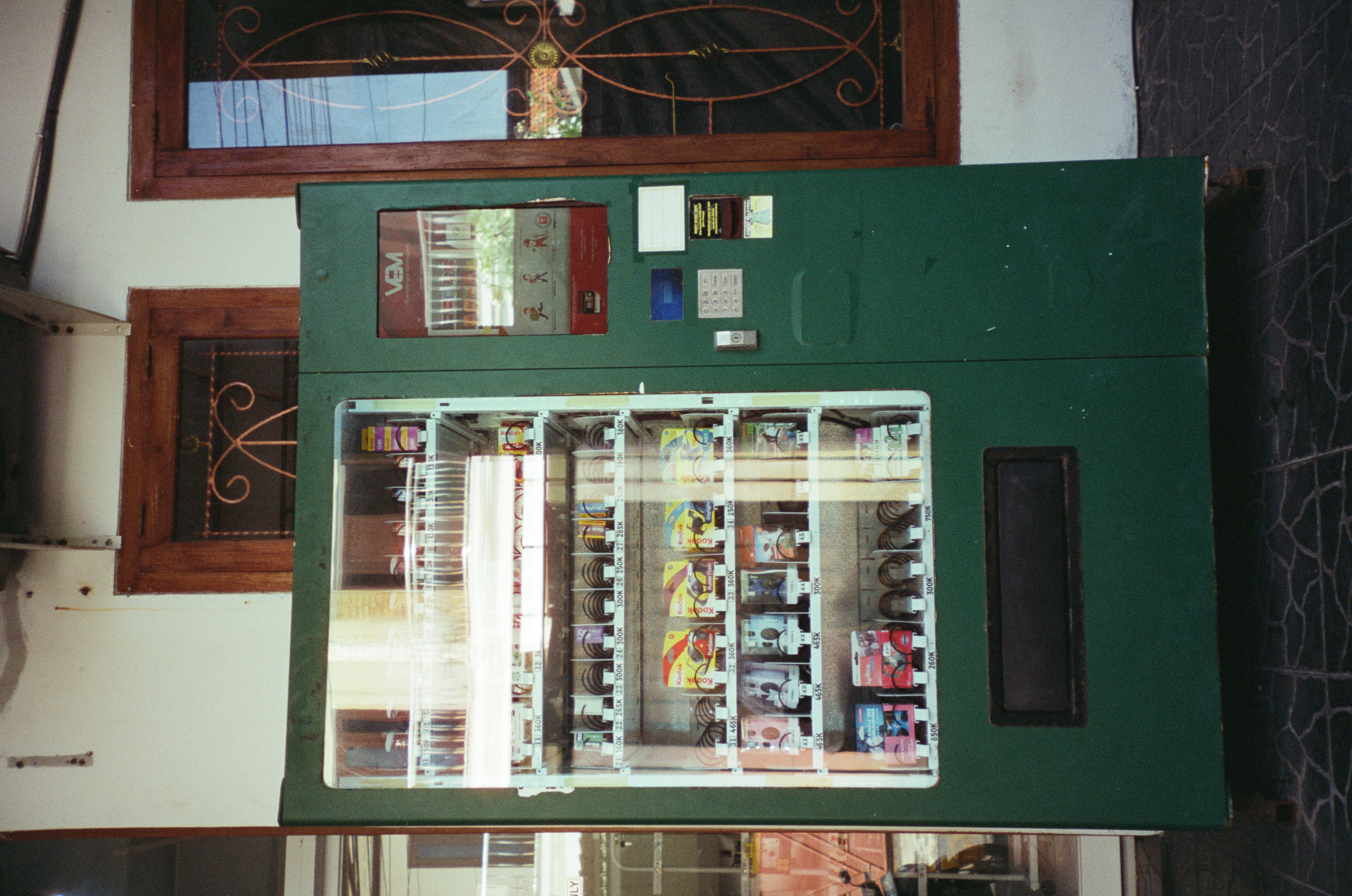 Weathered green vending machine with lit shelves of film canisters, boxed Kodak single-use cameras, and priced slots stands between wooden doorframes with curled iron scrollwork.