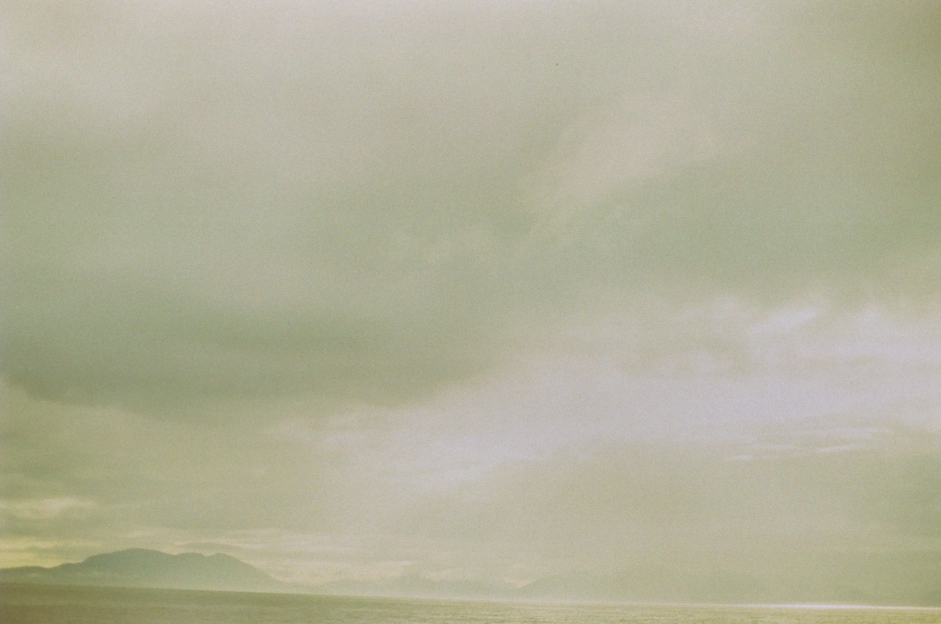 A dark low ridge stretches across pale water while gray clouds break into vertical bands of softened light.