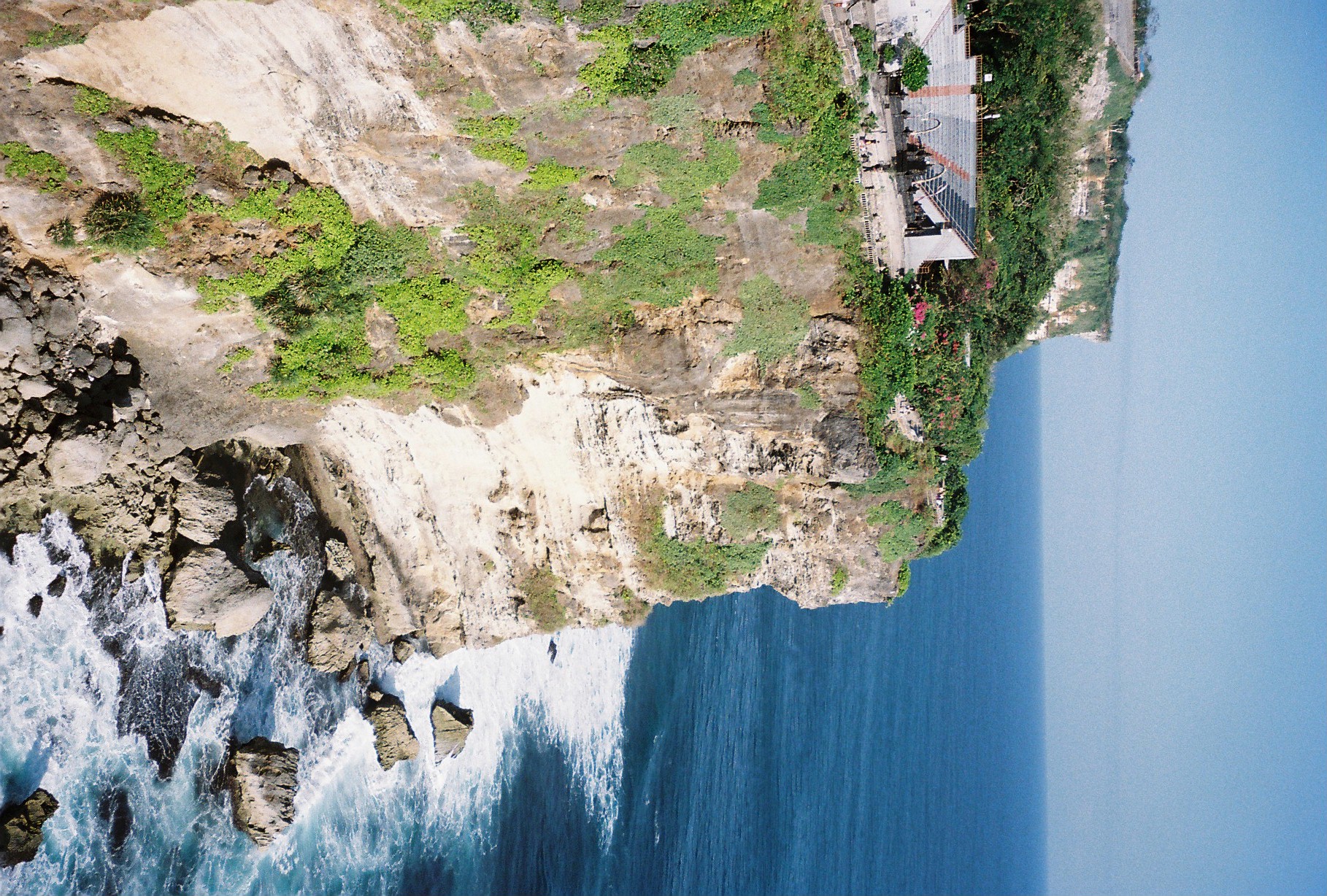 Layered rock strata run down a cliff face toward foaming surf and jagged rocks, with a small roofed structure perched at the clifftop among shrubs.