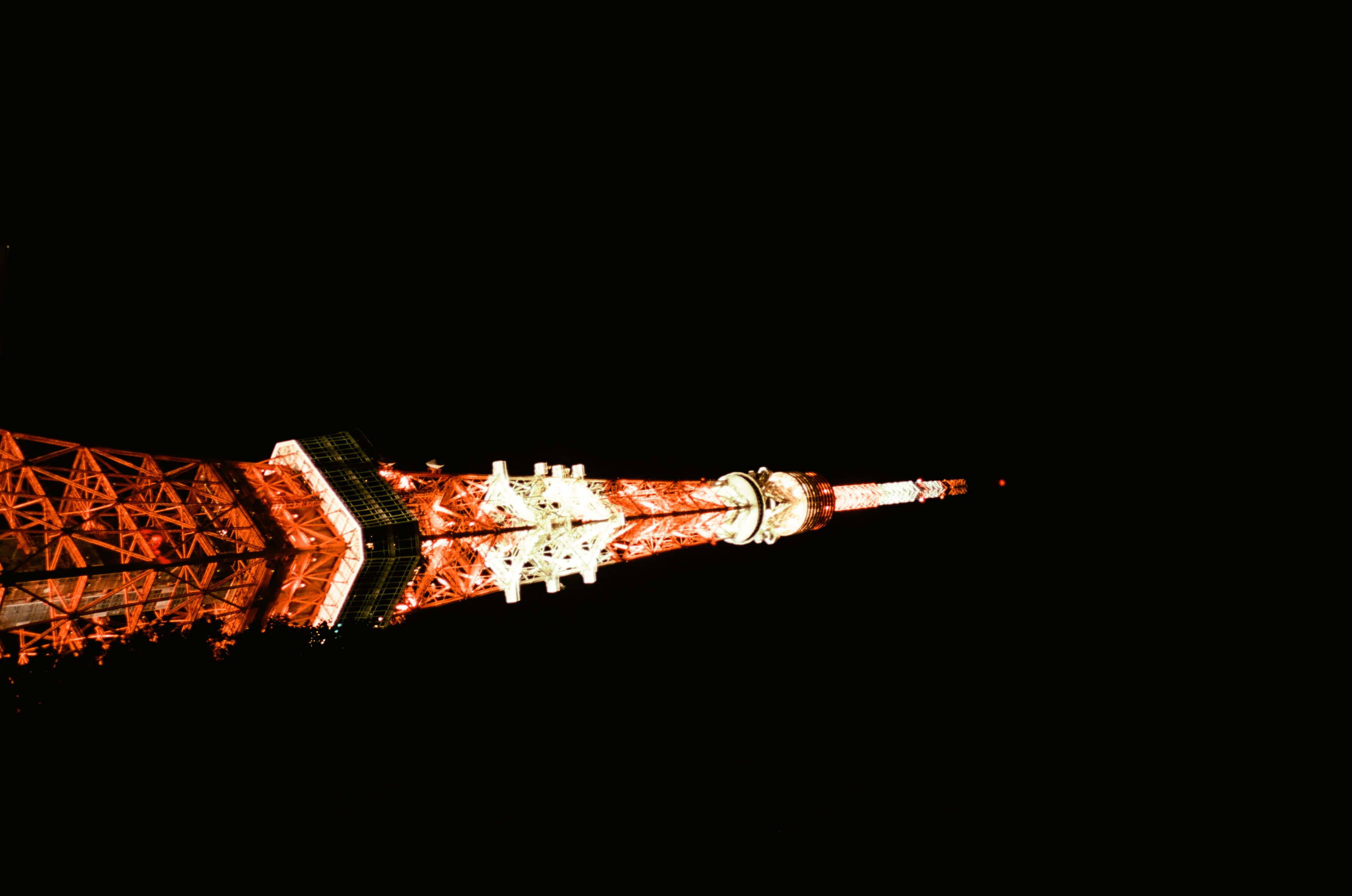 The white and orange antenna spire and upper platforms of a lattice tower float alone against a completely black sky.
