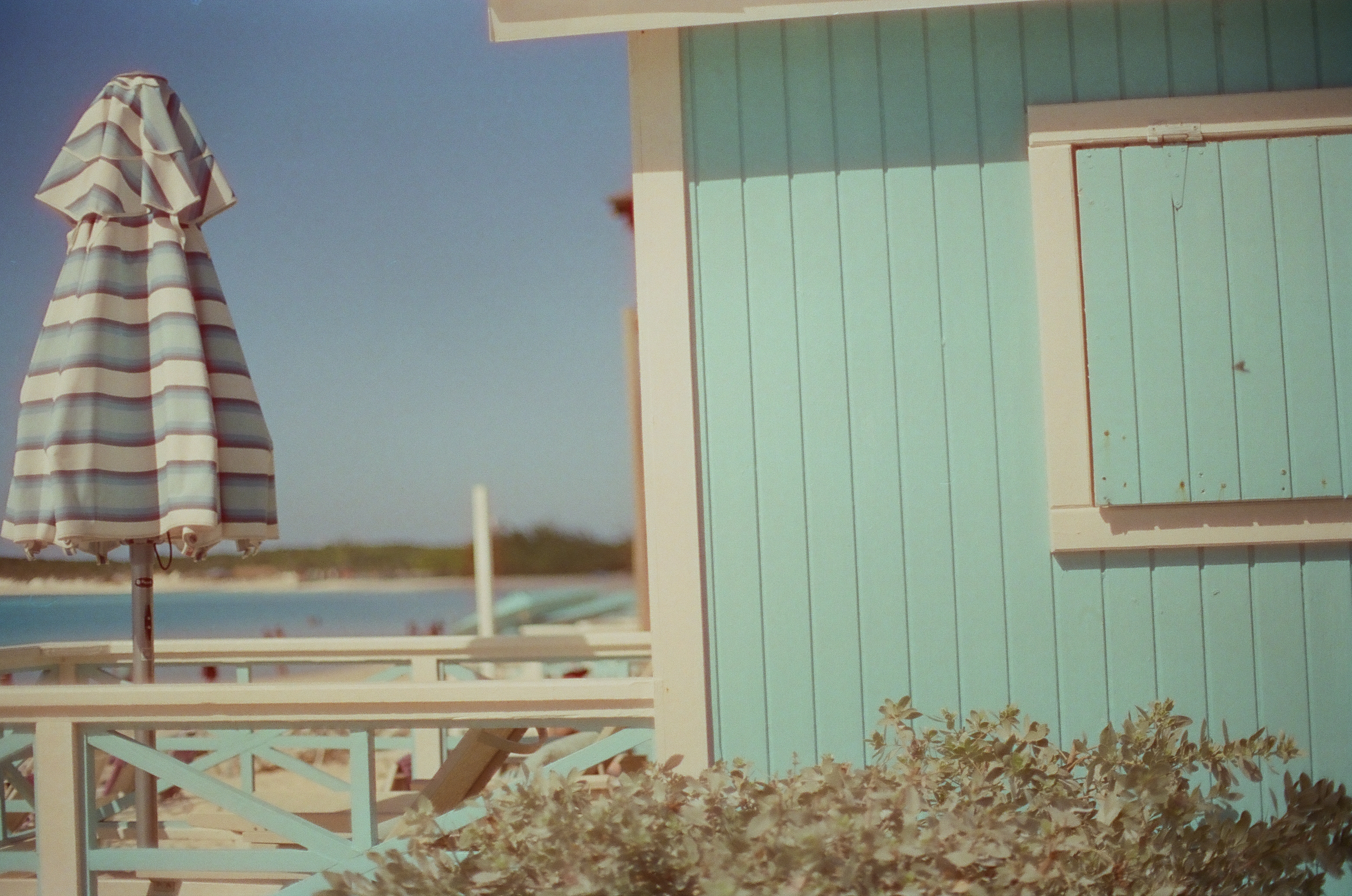 A closed striped umbrella stands beside a turquoise wooden wall and white railing with calm water beyond.