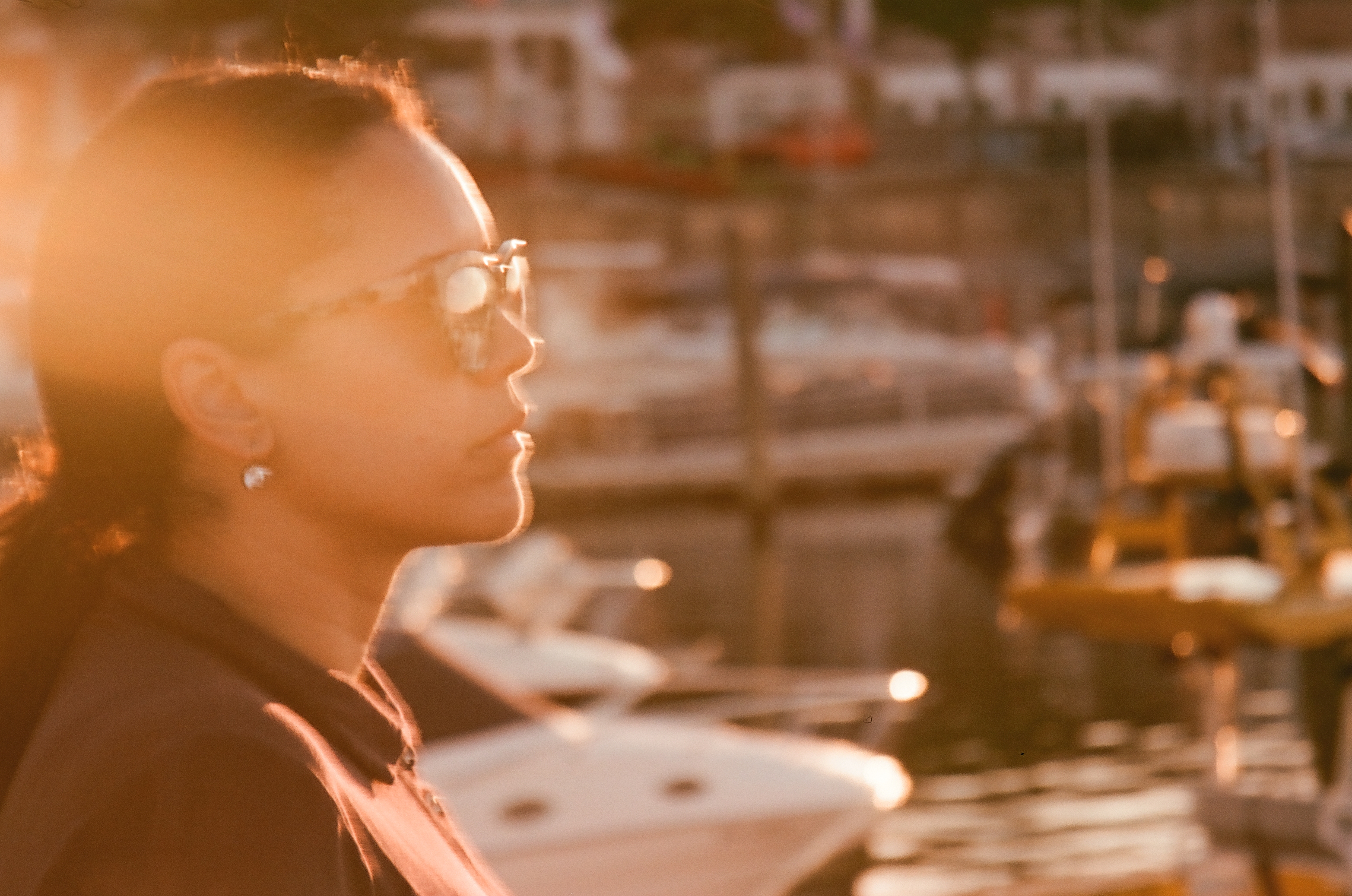 Woman in sunglasses in profile, backlit by low sun with strong orange flare, blurred masts and boats across the water behind her.