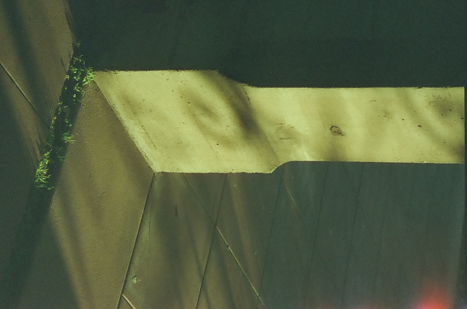 A narrow stucco column casts a long diagonal shadow across square outdoor tiles edged by a strip of grass.