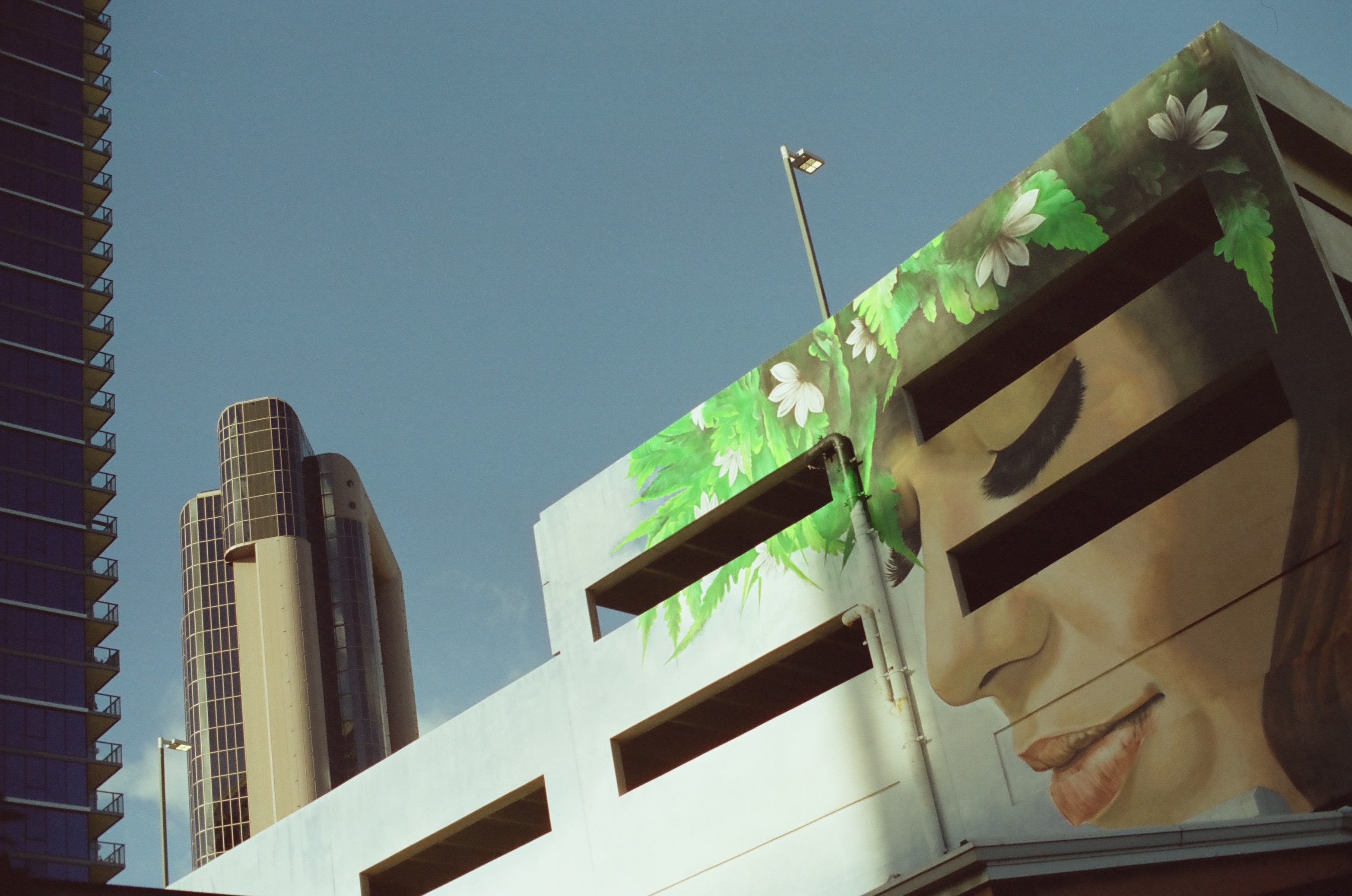 A large cropped face and green foliage cover a parking structure while a cylindrical high-rise stands beyond.