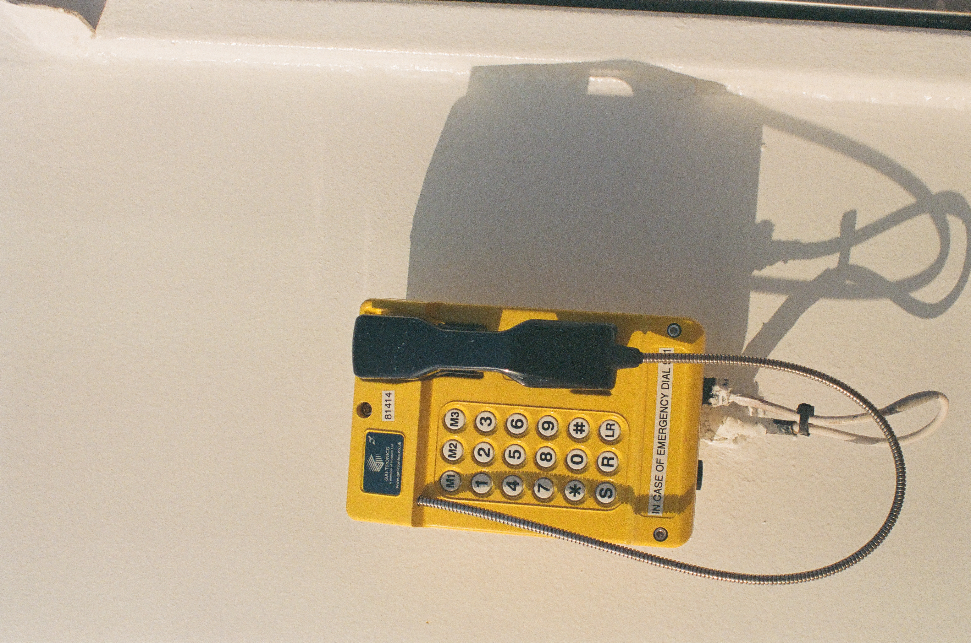 A yellow emergency telephone with a numbered keypad is mounted on a white wall, its handset and coiled cord casting a sharp shadow.