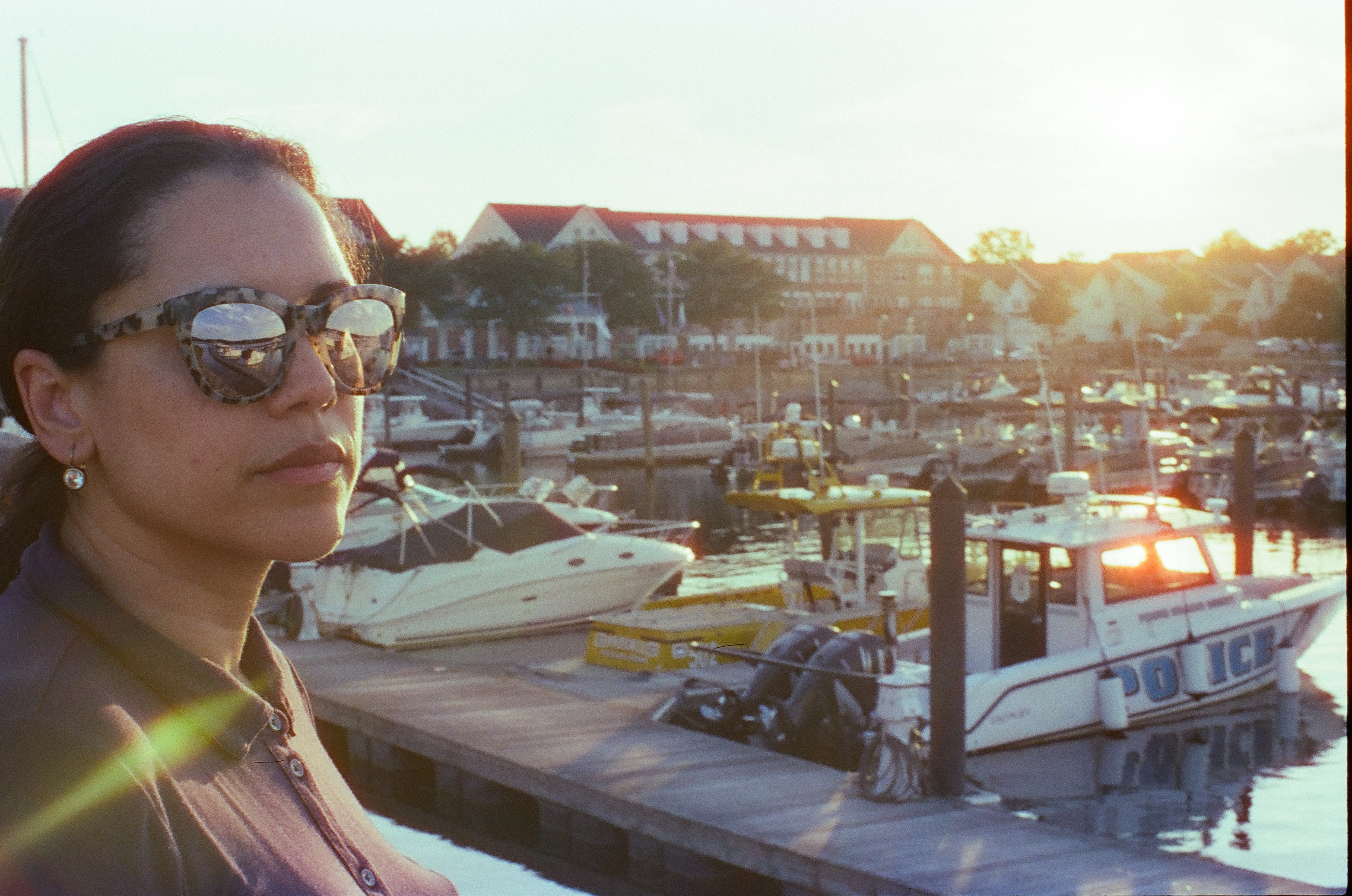 Woman in patterned sunglasses facing the camera on a dock, with moored powerboats, a white boat bearing partially hidden blue letters, and red-roofed buildings behind.