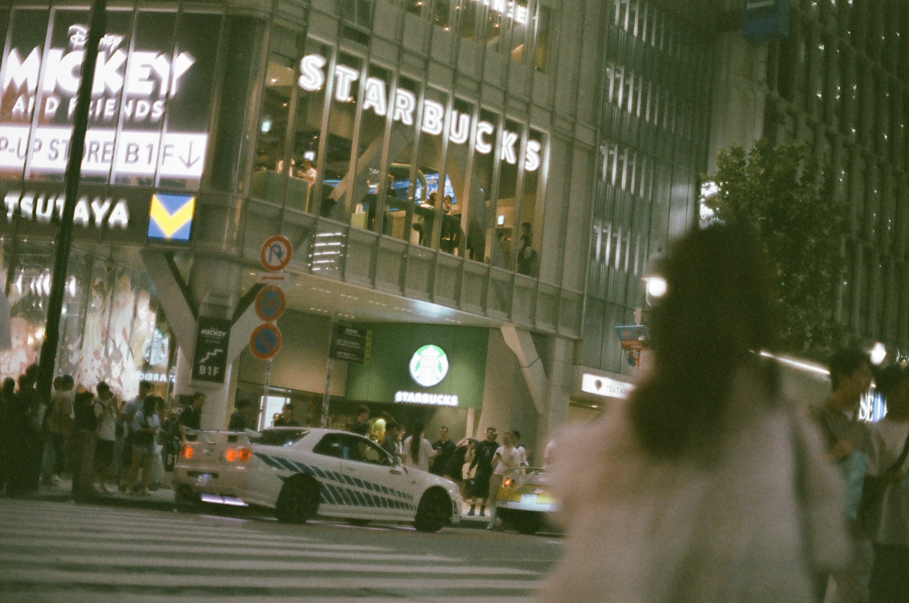 A white car with dark racing stripes rolls through an intersection past a lit corner marked 'STARBUCKS,' 'TSUTAYA,' and 'MICKEY AND FRIENDS.'