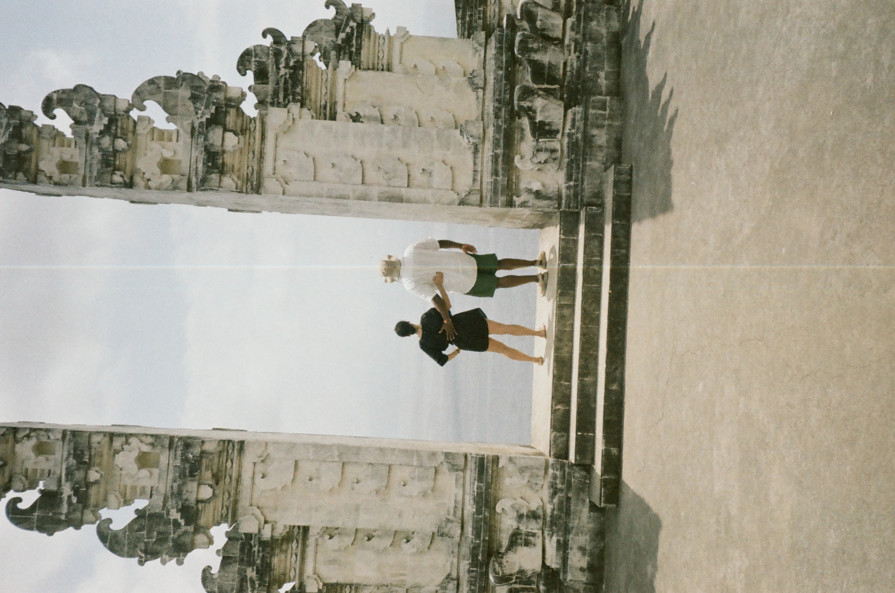 Two people seen from behind stand between carved towers and face a hazy blue-gray distance beneath cloudy sky.
