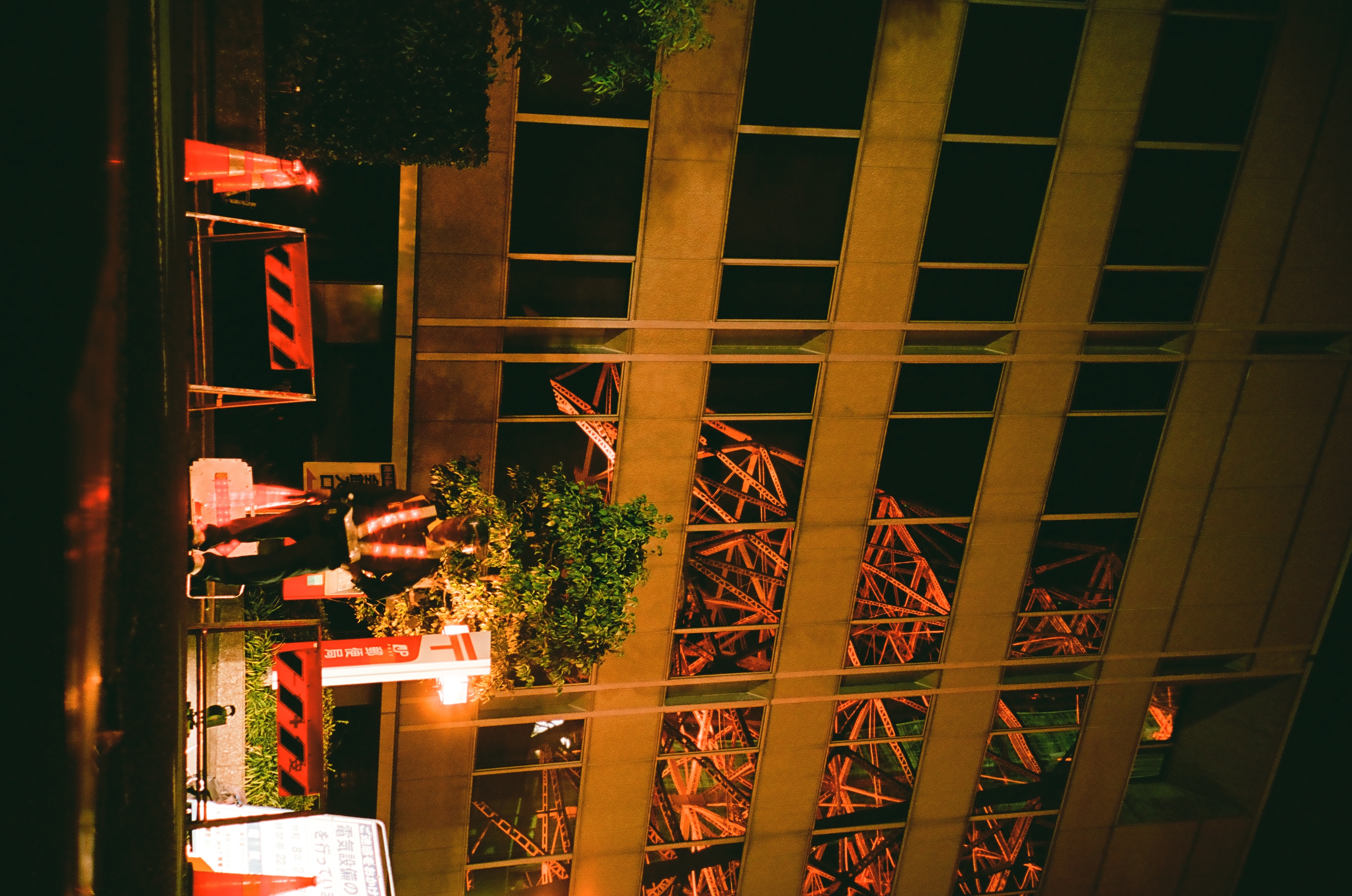 A dark gridded building catches red structural reflections above street barriers, small trees, and pools of warm night light.