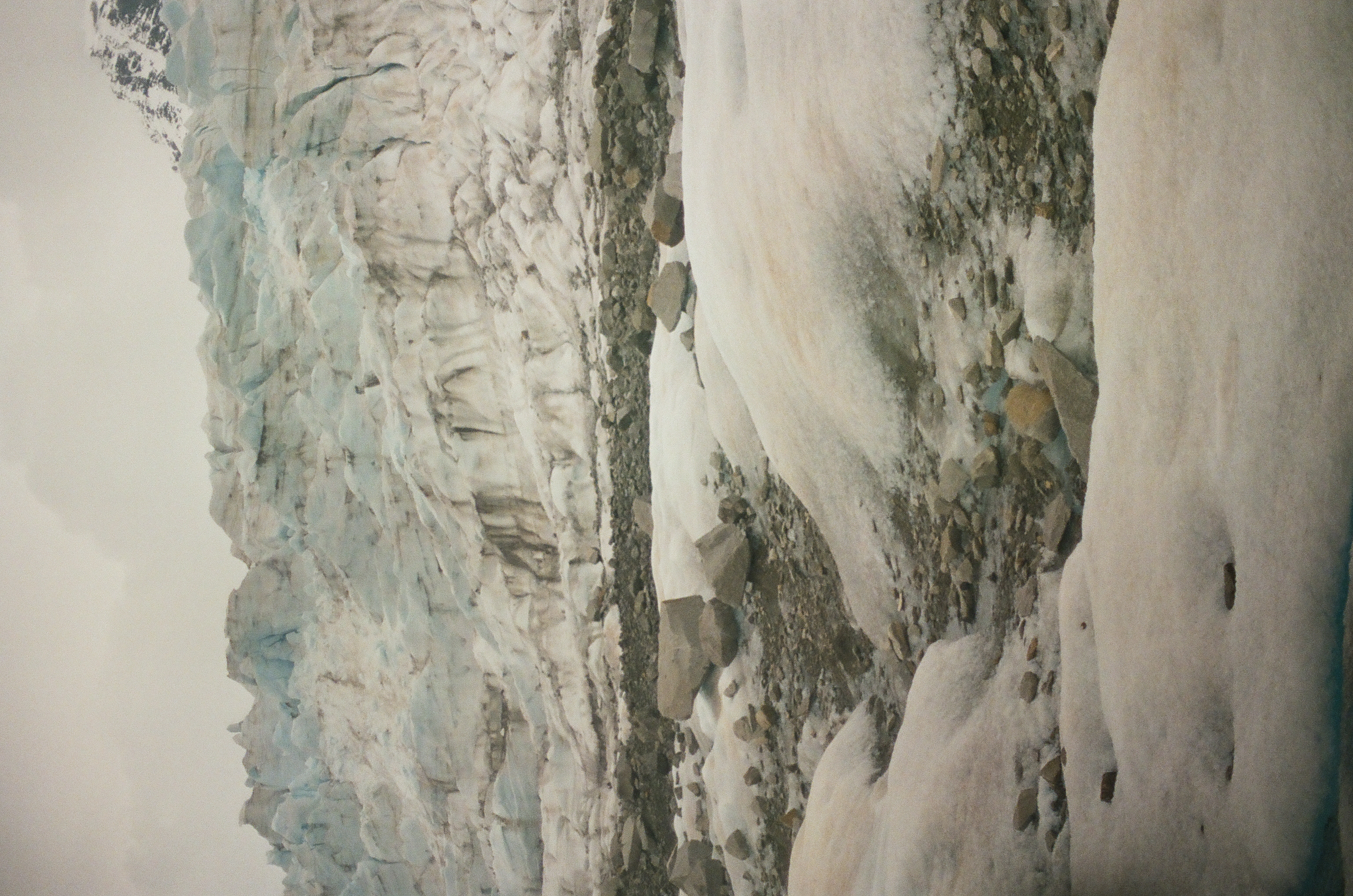 A deeply fractured glacier fills the foreground below distant dark peaks and an overcast white sky.
