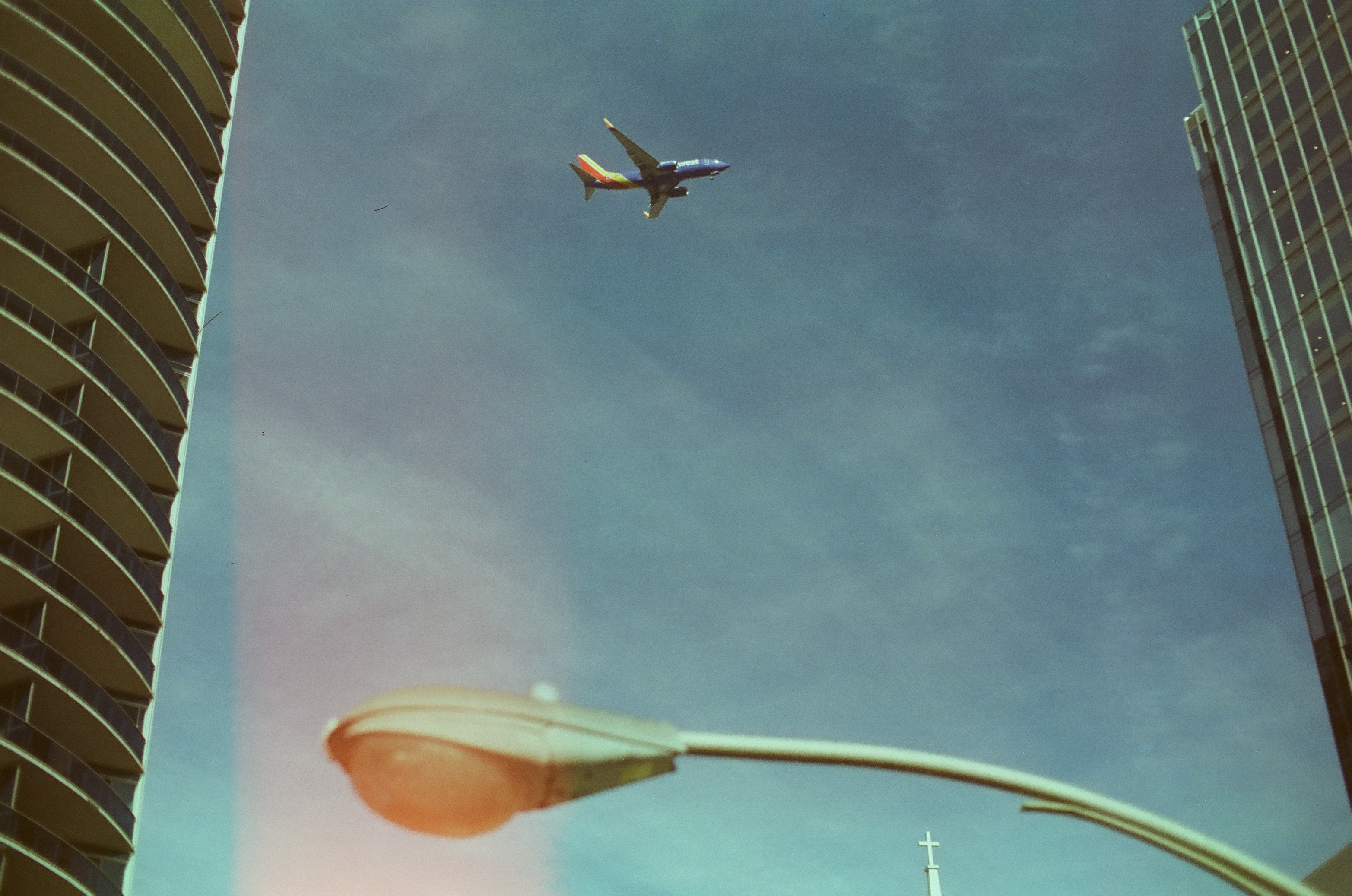 A jet crosses blue sky between a rounded balcony tower and a rectilinear glass building above a curved streetlight.