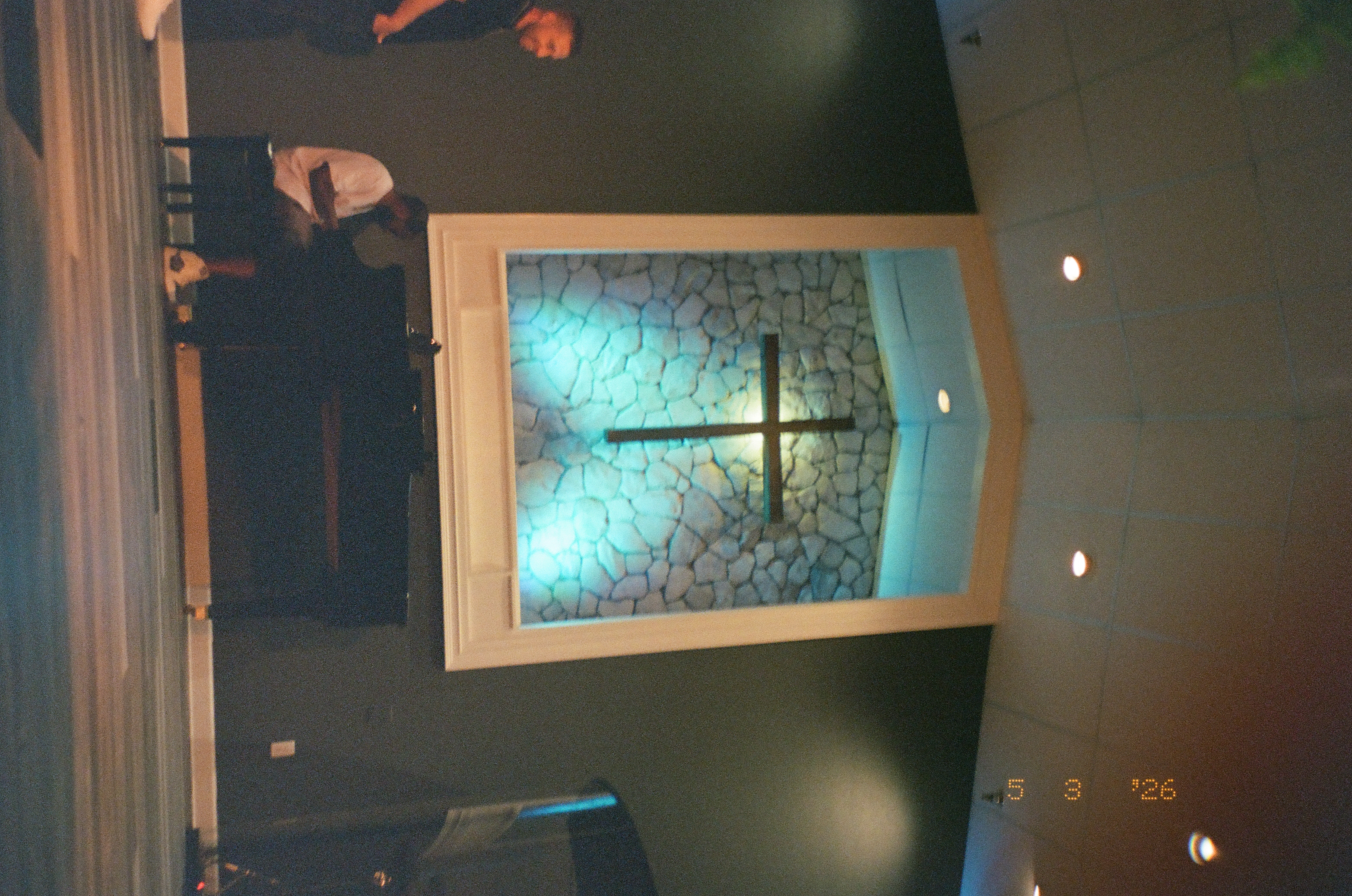 A wooden cross mounted on a stone-textured panel lit blue hangs above two men, one seated at an upright piano and one standing beside it.