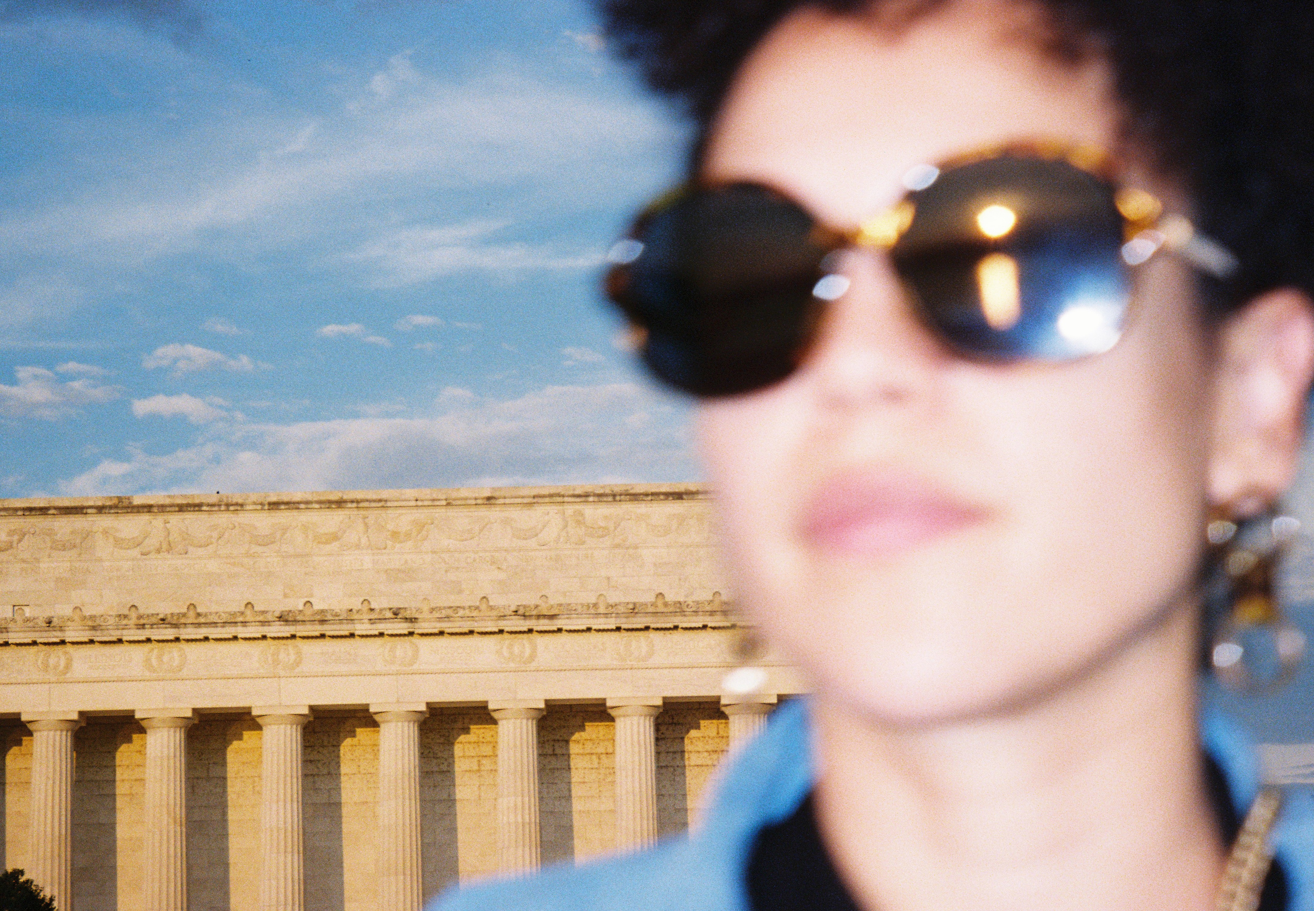A softly focused person wearing sunglasses stands before a long row of sunlit stone columns.