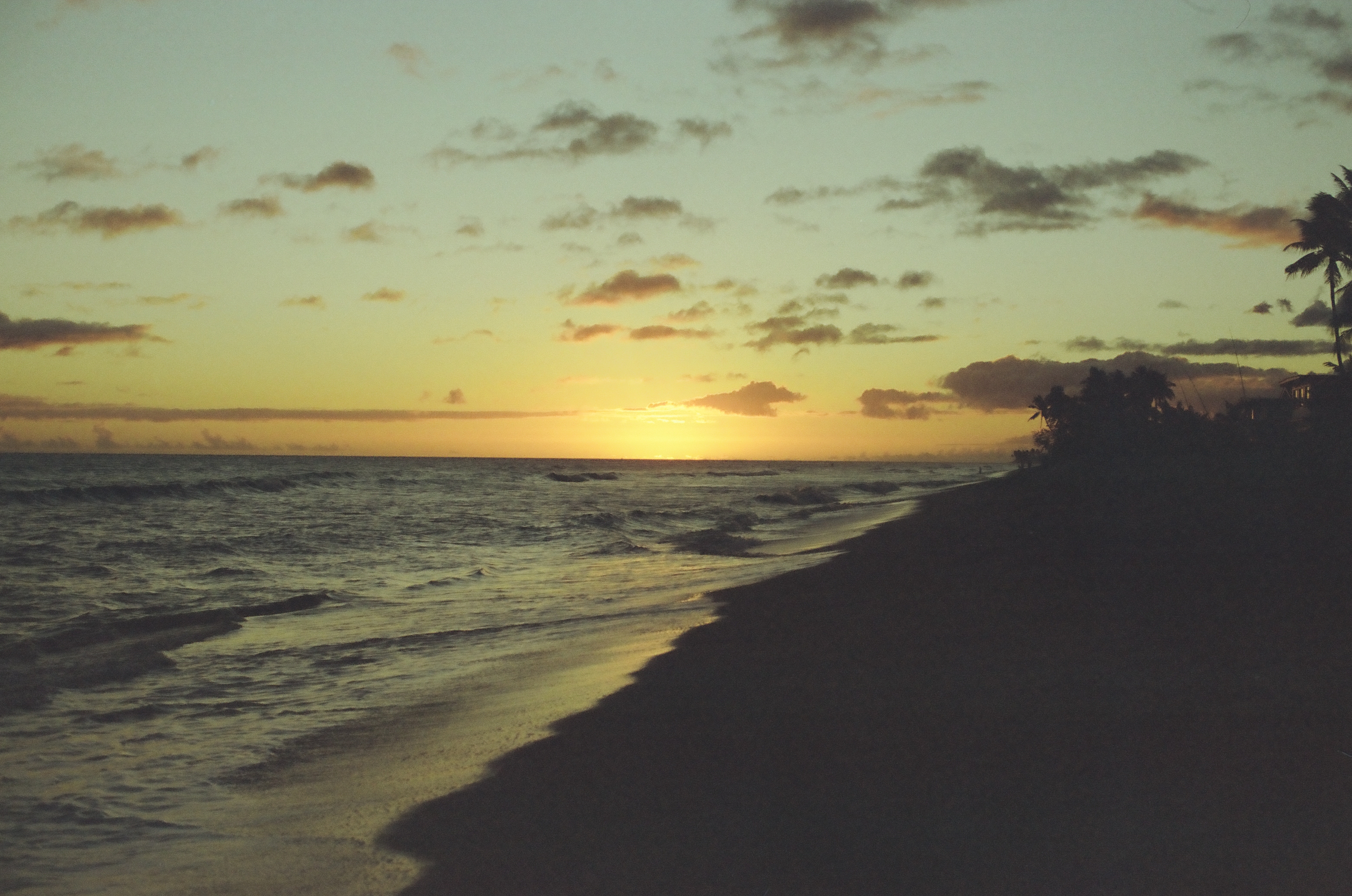 Golden sunset light stretches across the ocean beside a dark curving shoreline and scattered clouds.