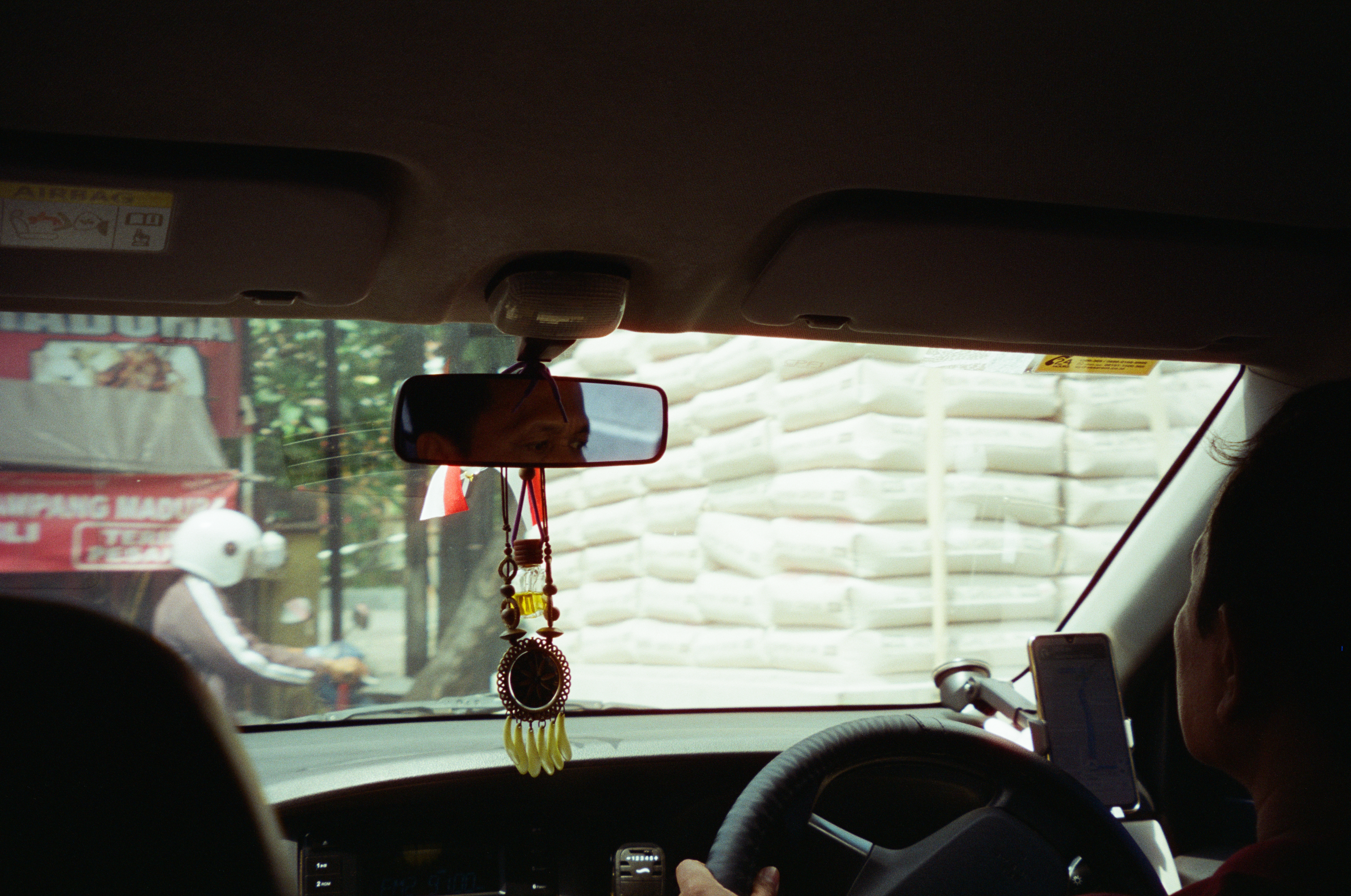 A charm of beads, a small bottle, pale tassels, and a red-and-white streamer hangs from a rearview mirror reflecting a driver's face, with a sack-loaded truck filling the windshield.