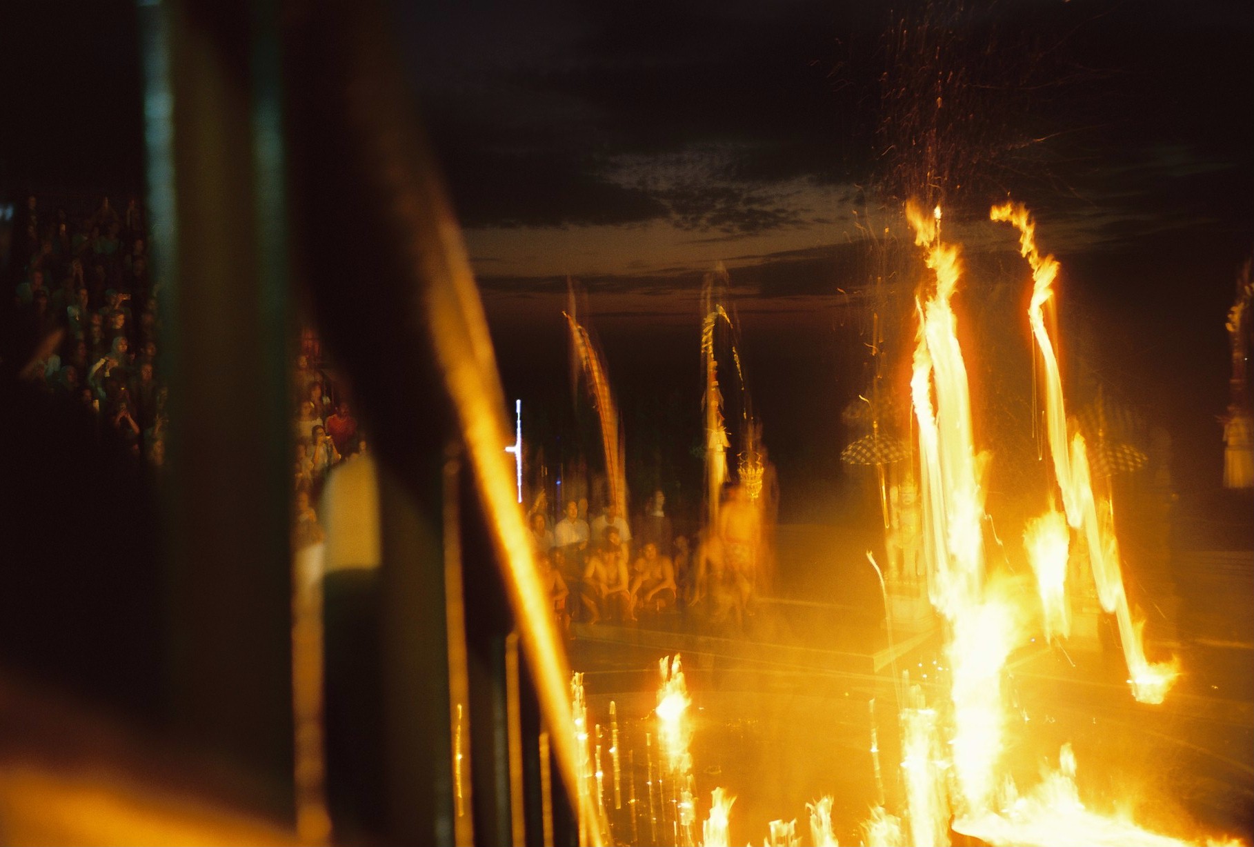 Two narrow columns of flame rise above a larger fire while rows of seated and standing spectators watch beyond.