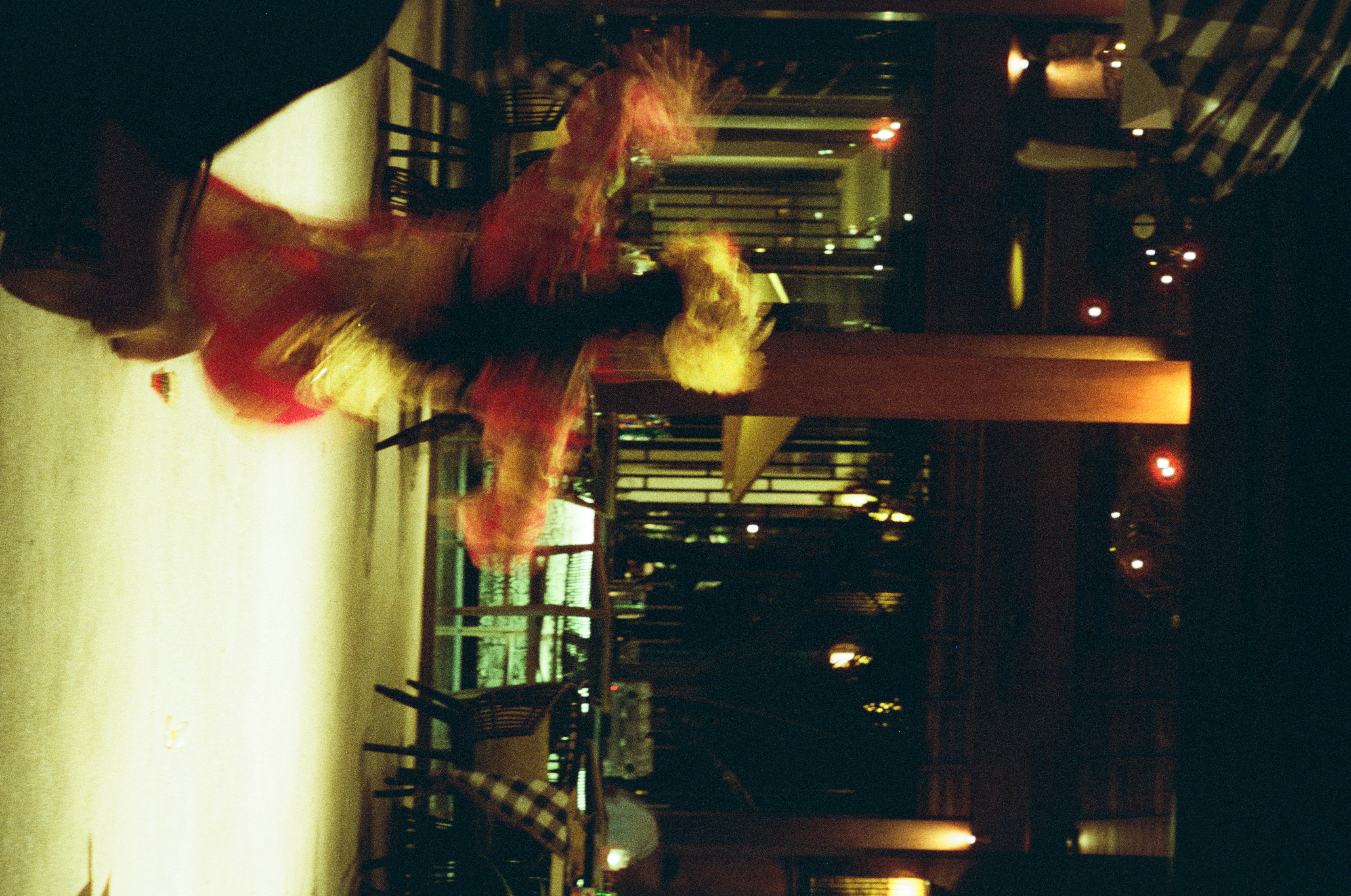 A red-and-gold costumed figure with a large gold head adornment extends blurred arms across a bright floor, with tables, columns, lamps, and checked fabric behind.