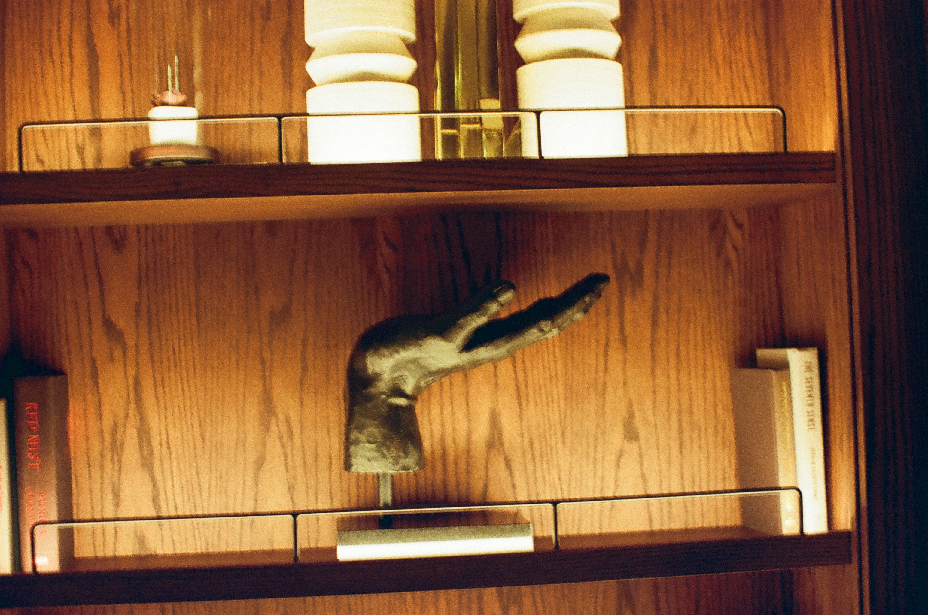 A dark bronze hand sculpture reaches upward from a wood shelf beside paperback books, below fluted lamp bases on the shelf above.
