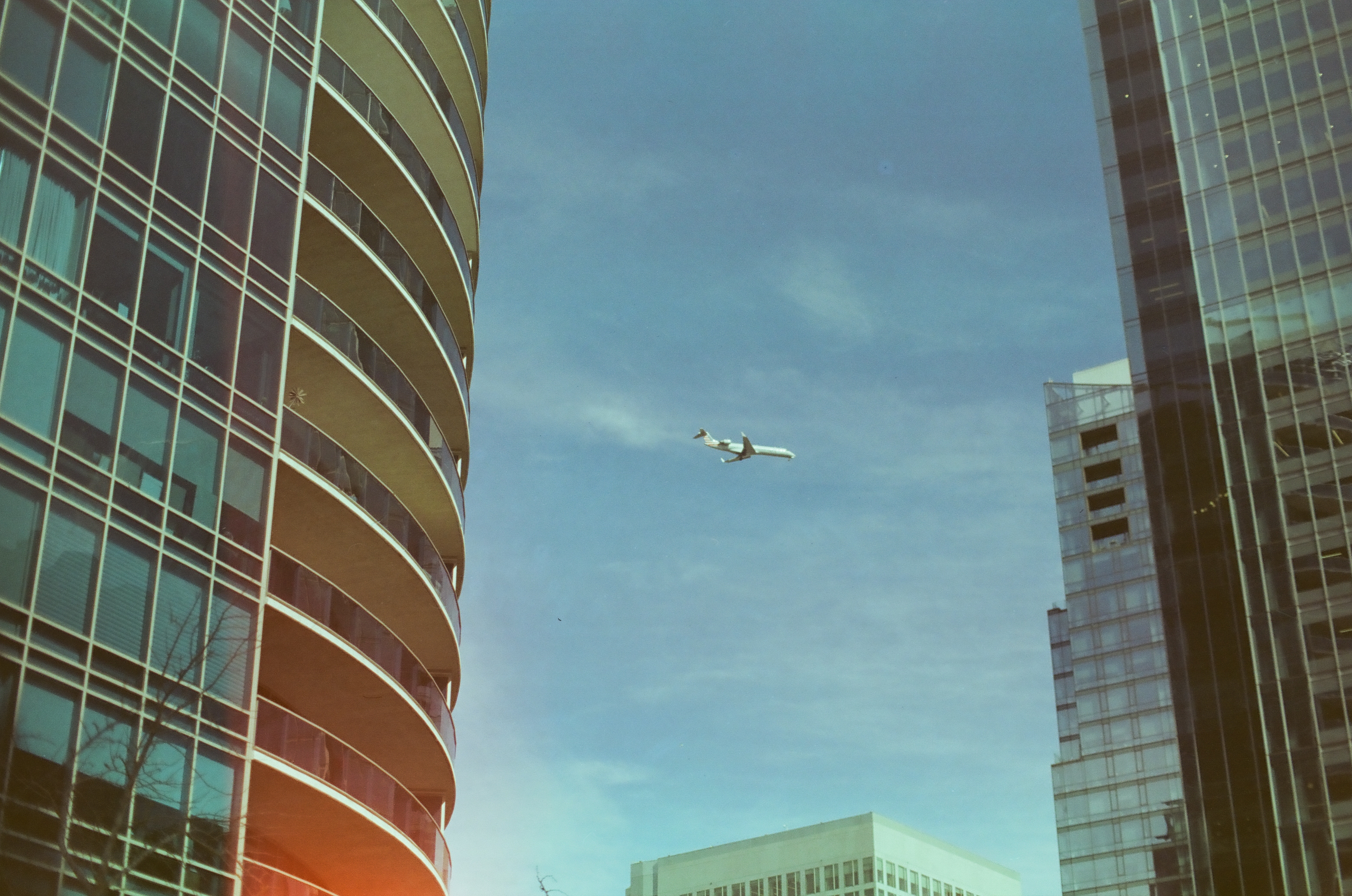 A small aircraft appears in open blue sky between curved glass balconies and tall reflective rectangular buildings.