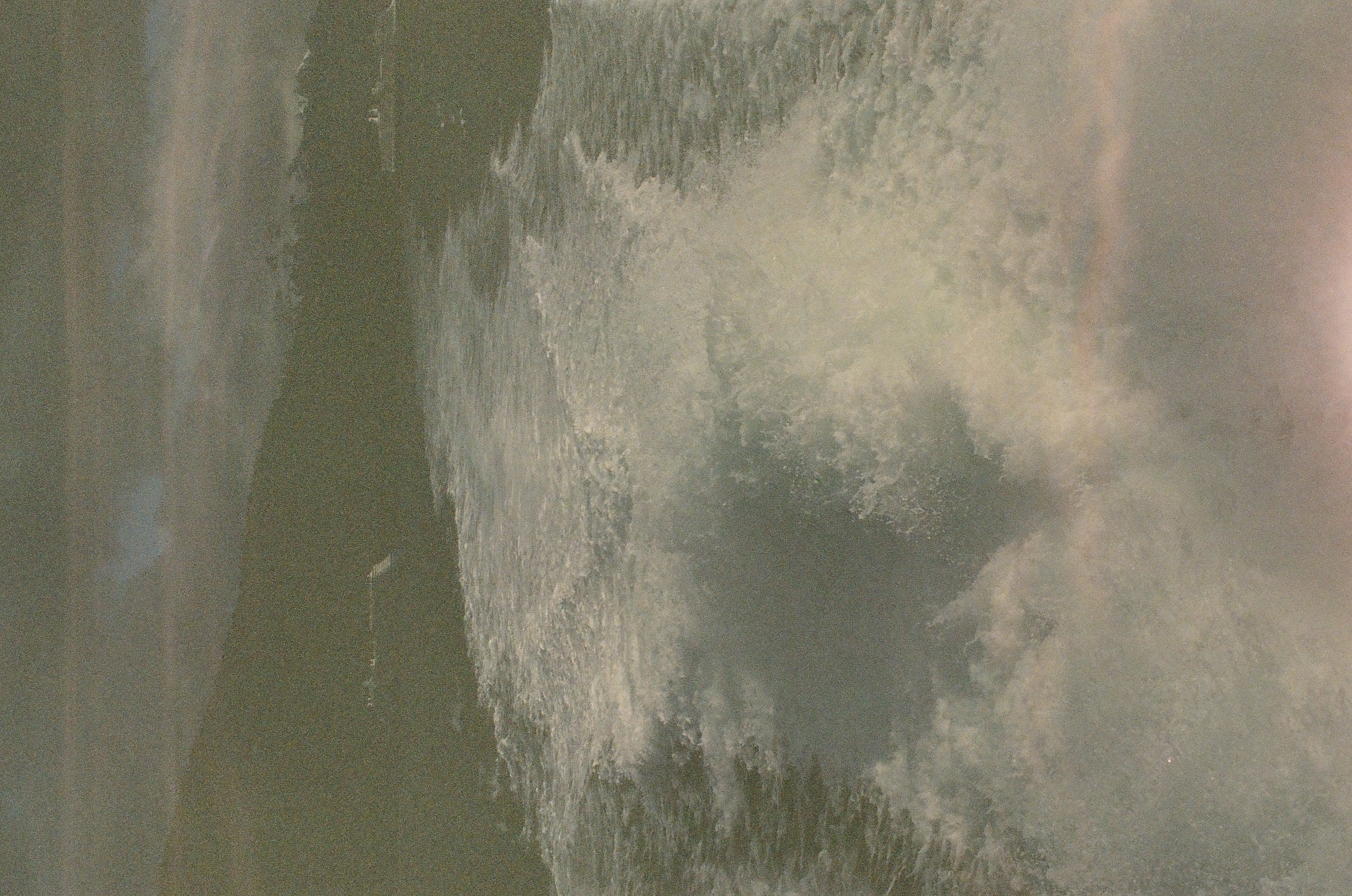 A broad white wake crosses dark water toward a forested shoreline and low clouded mountains.