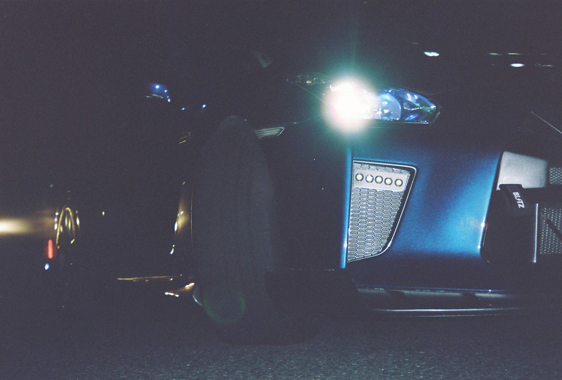 Ground-level view of a blue car's front corner with one headlight glaring, a mesh vent with five small lights, and dark pavement.