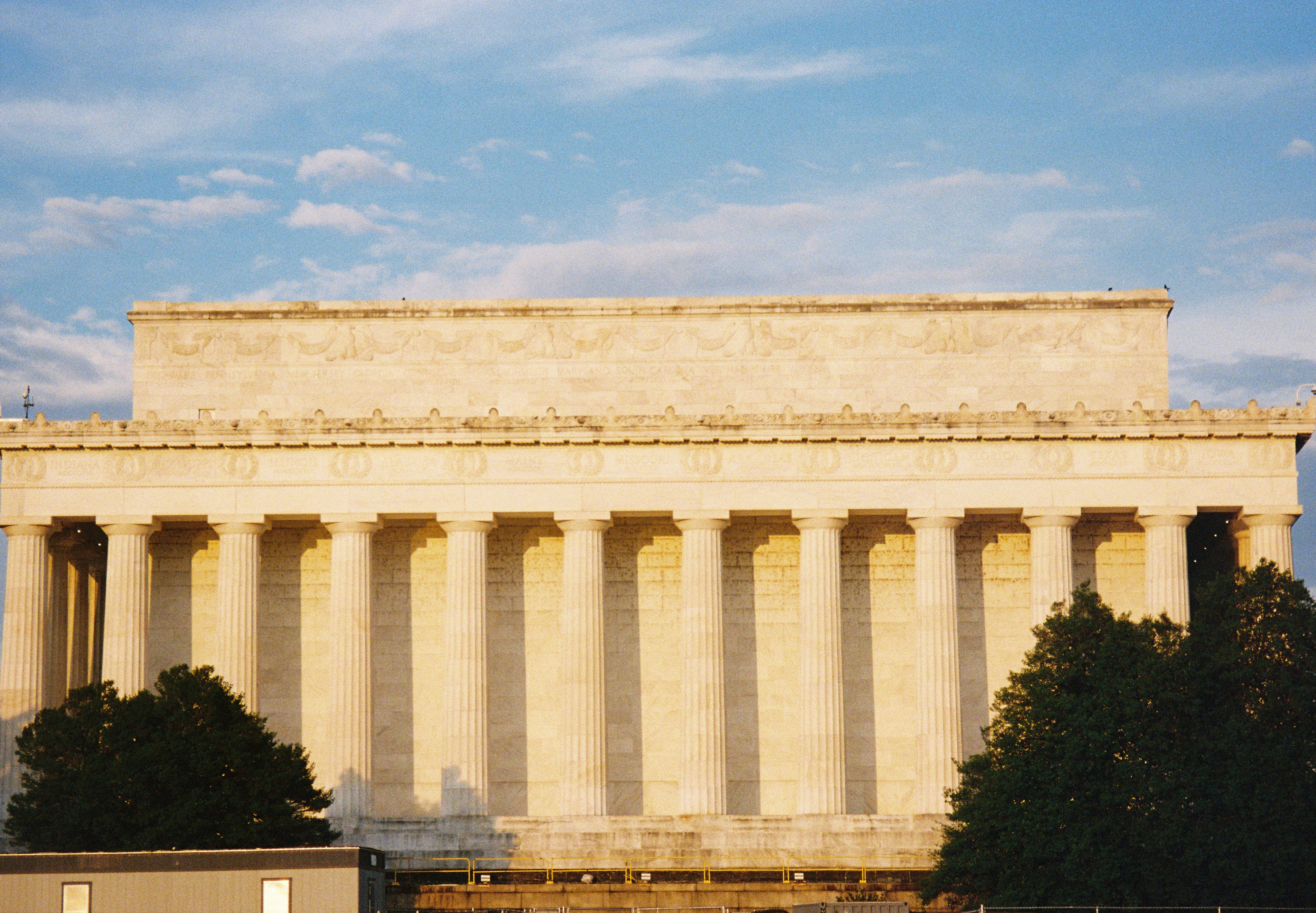 A rectangular memorial with a long colonnade stands between dark trees and a softly clouded blue sky.
